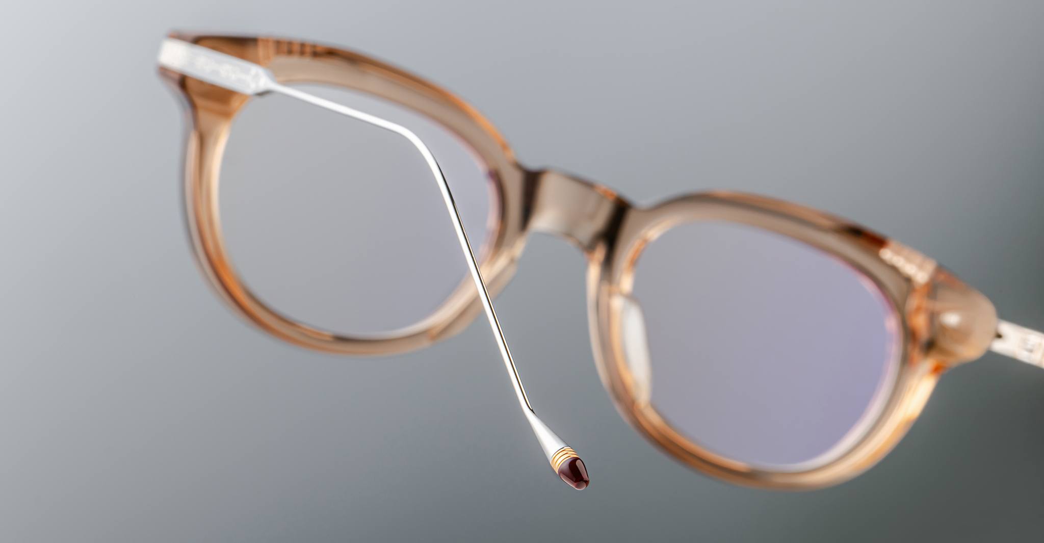 A close-up of the Jacques Marie Mage Hisao eyeglasses featuring light brown, translucent round frames, highlighting the end of one temple arm against a neutral grey background.