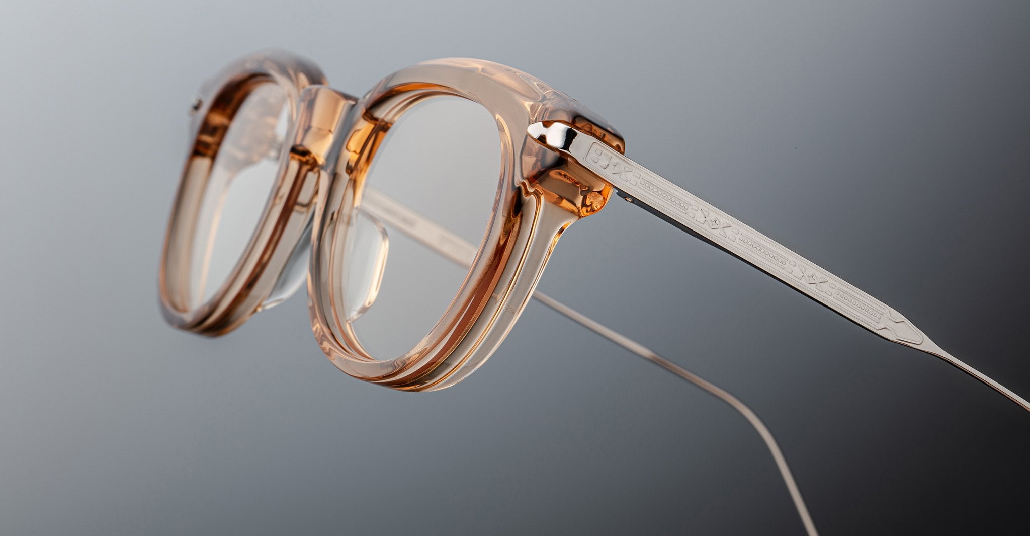 Close-up of Jacques Marie Mage Hisao eyeglasses featuring clear, thick round lenses and transparent peach-hued frames. The metal arms have engraved detailing, connect with visible screws, and the background is gray.