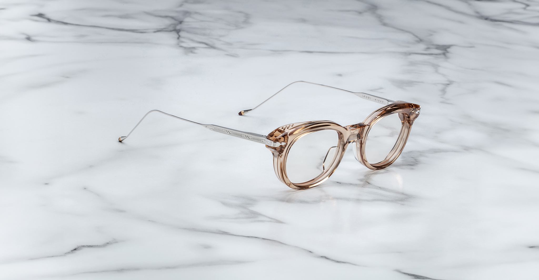 The Jacques Marie Mage Hisao eyeglasses, featuring a round shape in light brown with silver metal arms, rest on a white marble surface with gray veining.