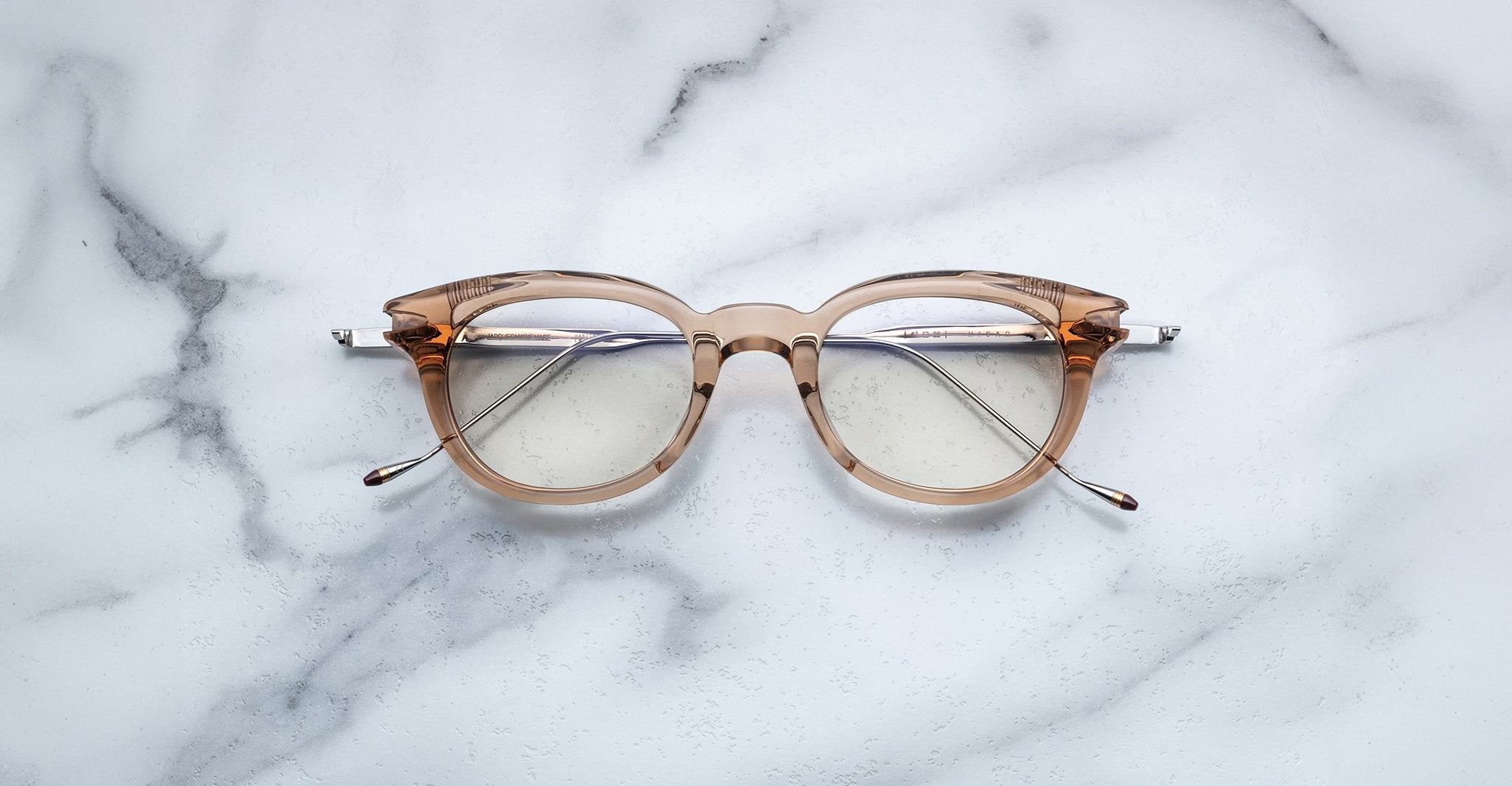 A pair of Jacques Marie Mage Hisao clear brown eyeglasses with silver temples rests on a white marble surface, front facing up and arms slightly open.