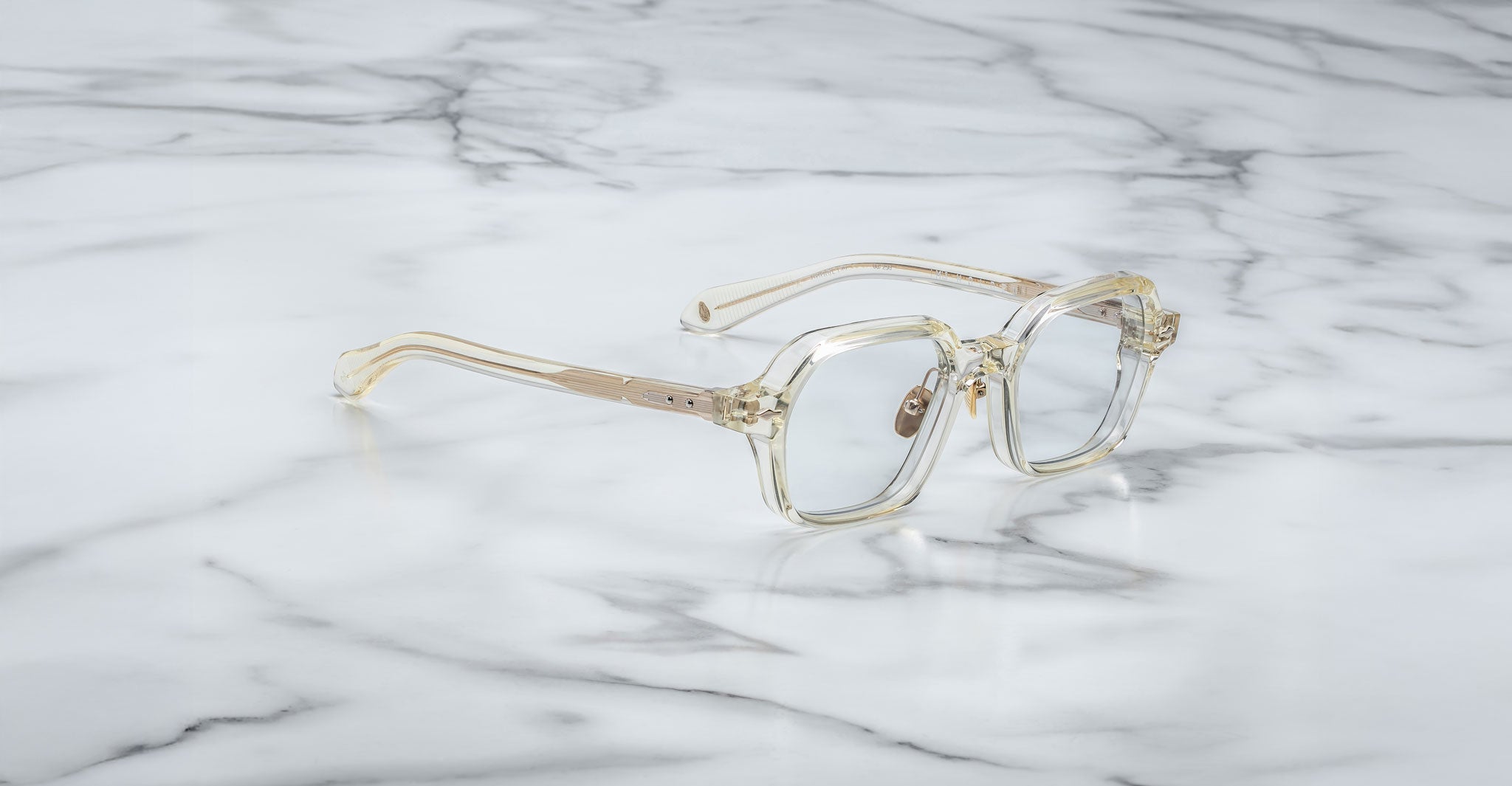 The Jacques Marie Mage Holden eyeglasses, featuring clear rectangular frames with gold accents, are displayed on a white marble surface with gray veining.