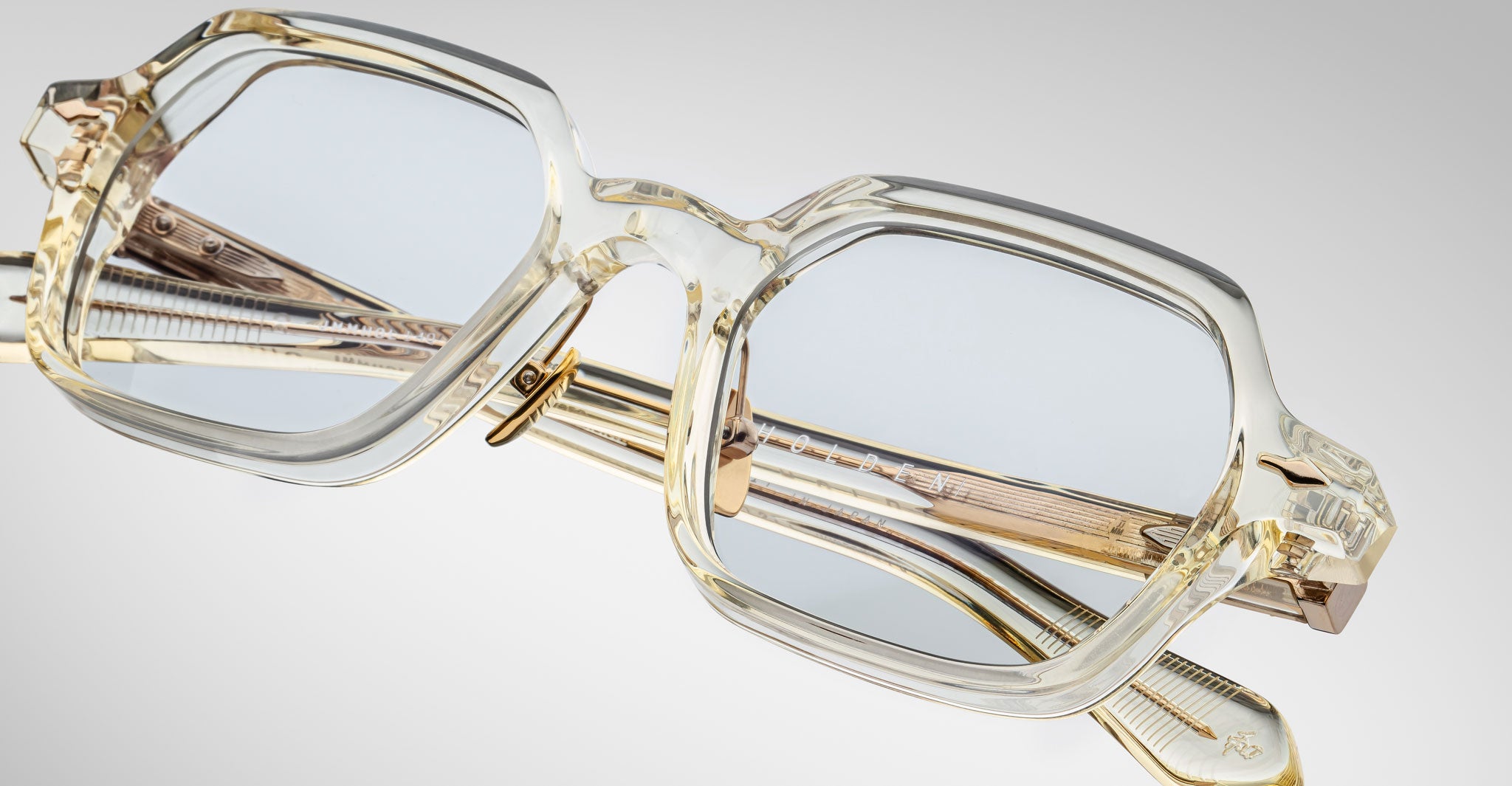 Close-up of Jacques Marie Mage Holden eyeglasses featuring stylish rectangular transparent pale frames and metallic gold arms, resting on a light gray surface. The lenses have a subtle blue tint.