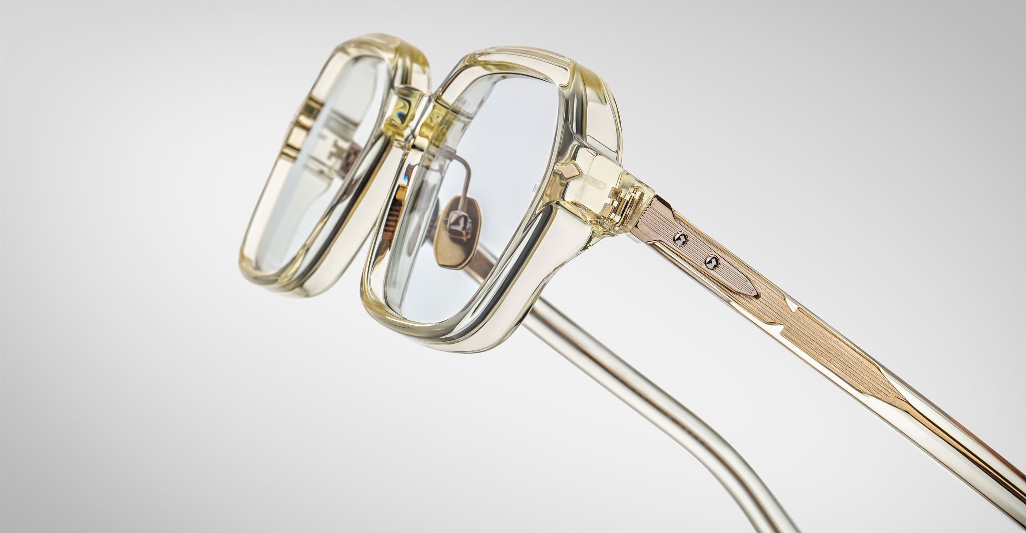 Close-up of the Jacques Marie Mage Holden eyeglasses featuring stylish, translucent clear frames and gold metallic temples, set against a light gray background. The glossy texture and modern design are prominently showcased.