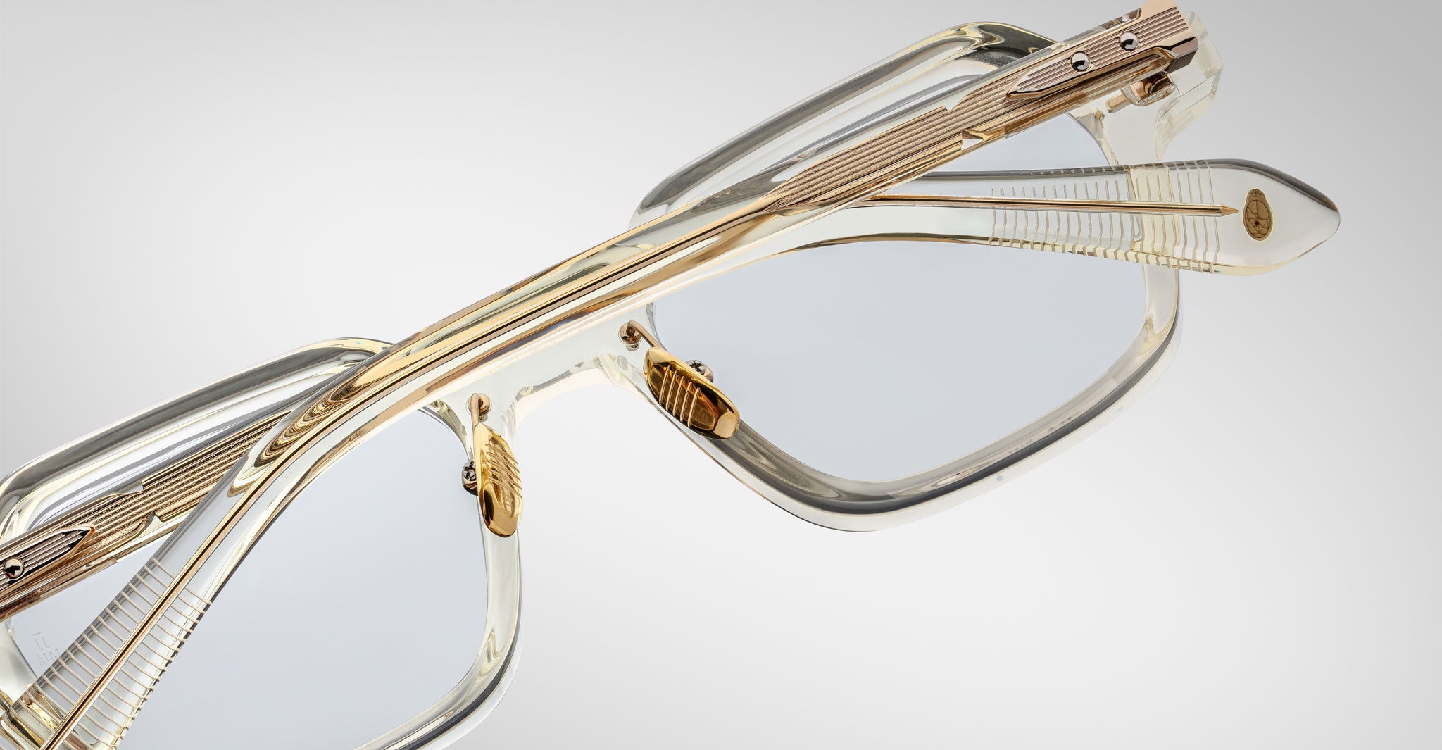 Close-up of Jacques Marie Mage Holden rectangular eyeglasses featuring transparent frames and gold accents on the arms and bridge, set against a light gray background.