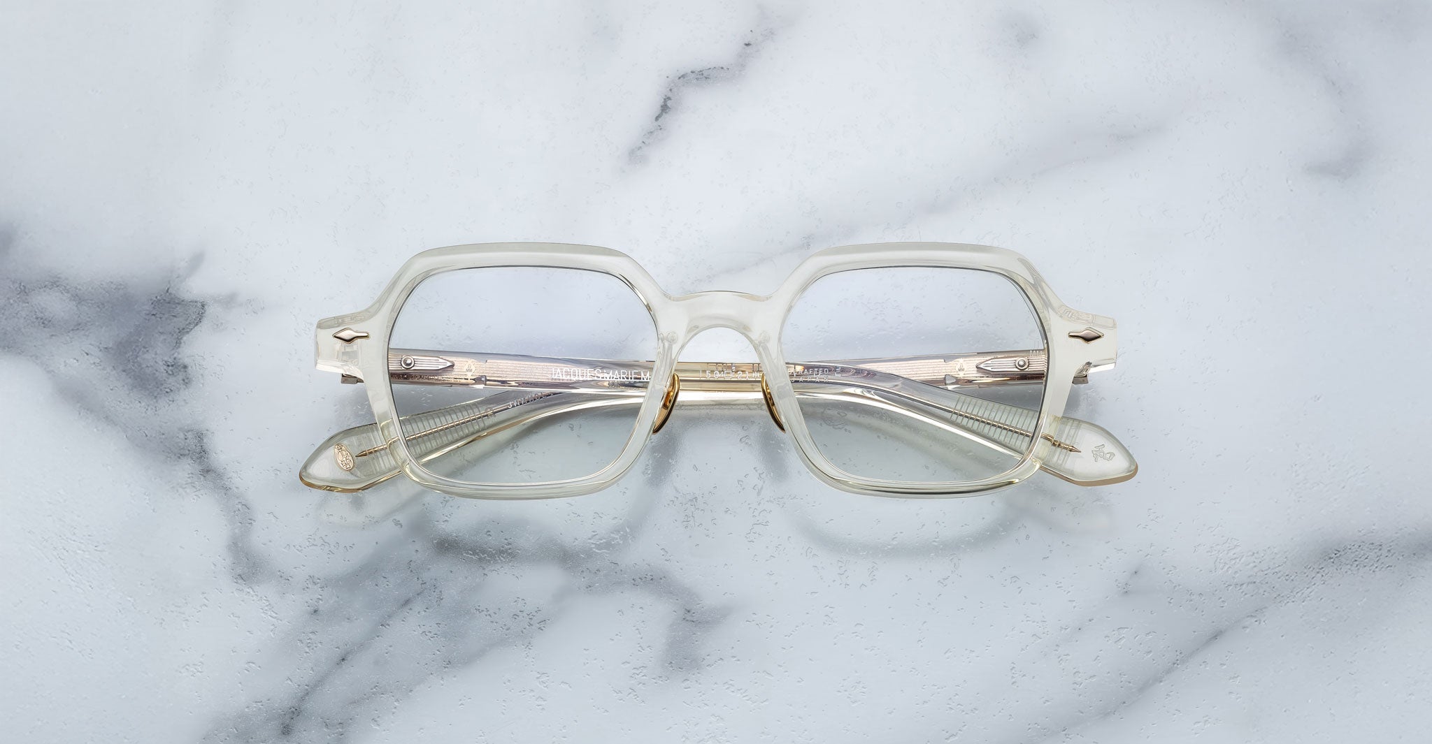 The Jacques Marie Mage Holden eyeglasses, featuring clear, square-shaped translucent frames, are displayed on a white and gray marble surface.