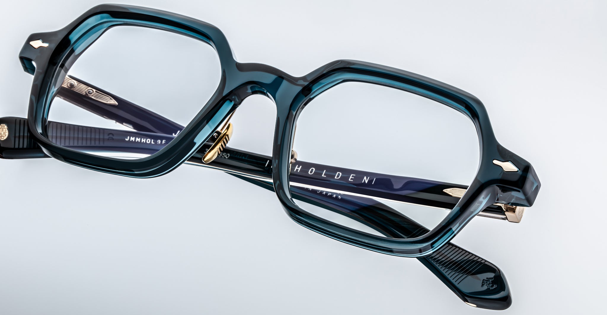 The Jacques Marie Mage Holden features stylish, thick-rimmed rectangular eyeglasses with dark teal frames and gold accents, resting on a white surface. The brand name “HOLDEN” is visible on the arm of the glasses.