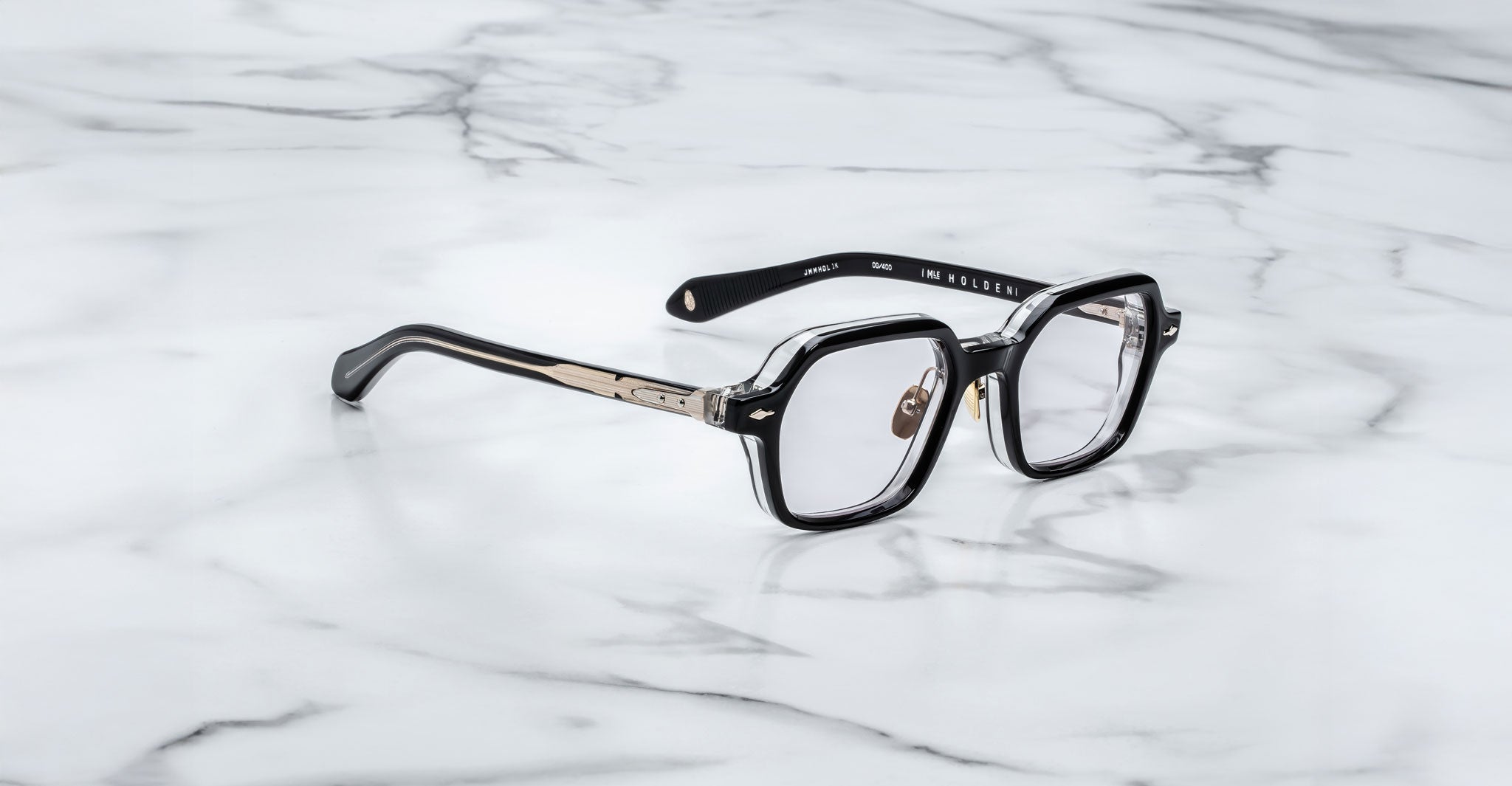 The Jacques Marie Mage Holden eyeglasses feature black and clear acetate geometric frames with metal-accented temples, shown on a white marble surface.