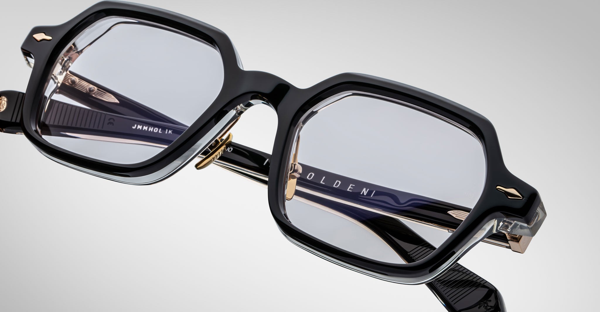Close-up of black rectangular Jacques Marie Mage Holden eyeglasses with gold detailing and clear corner accents, featuring the brand name on the inside of the temple against a light gray background.