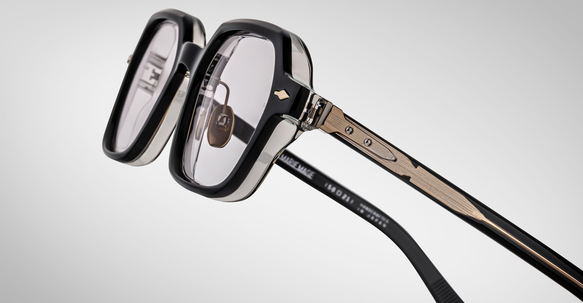 Close-up of Jacques Marie Mage Holden prescription eyeglasses featuring thick transparent square frames, black accents, gold metallic arms, and geometric detailing, set against a light gradient background.