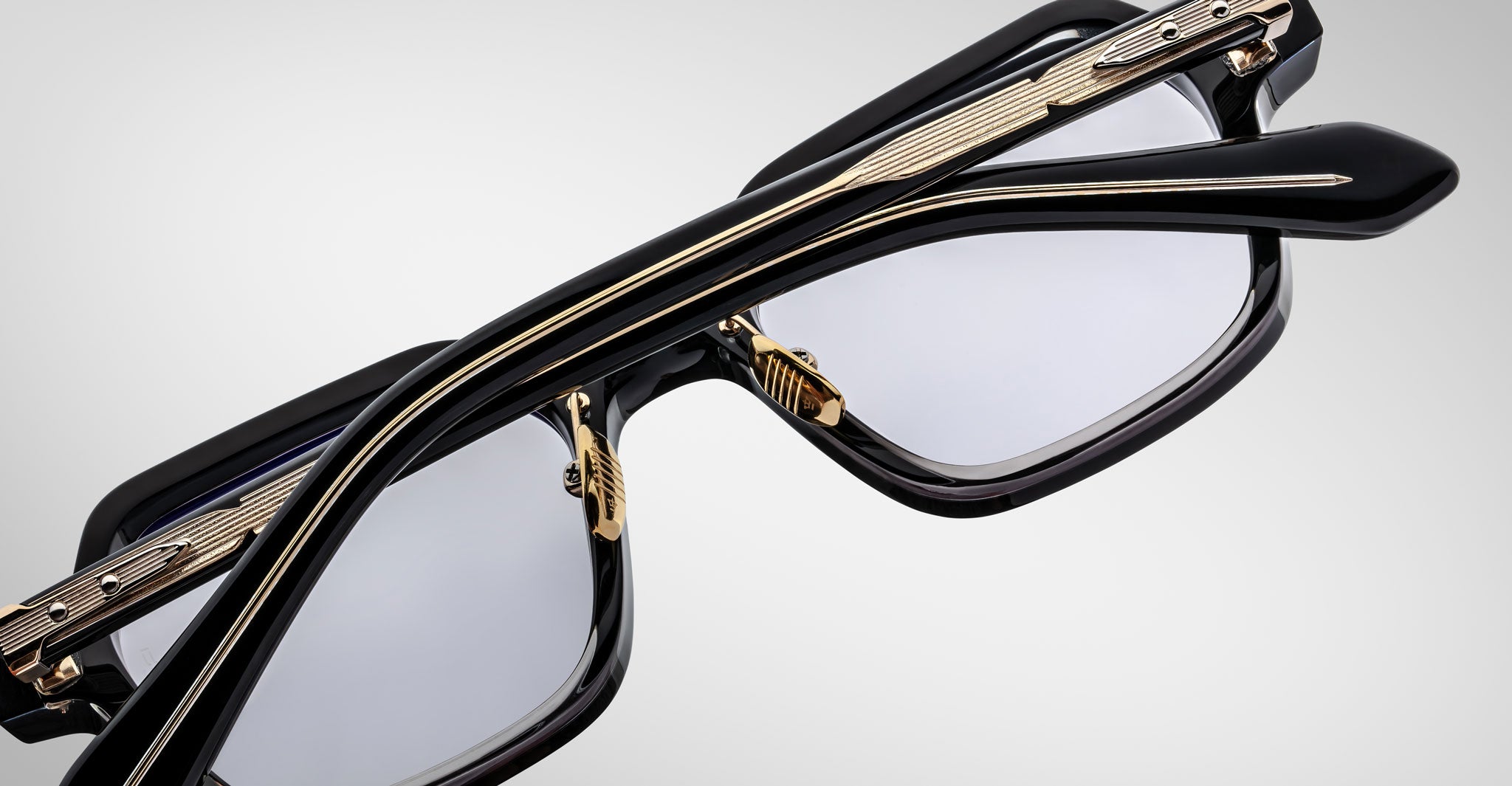 Close-up of two pairs of Jacques Marie Mage Holden eyeglasses, featuring black frames with gold detailing, stacked diagonally on a plain light background.