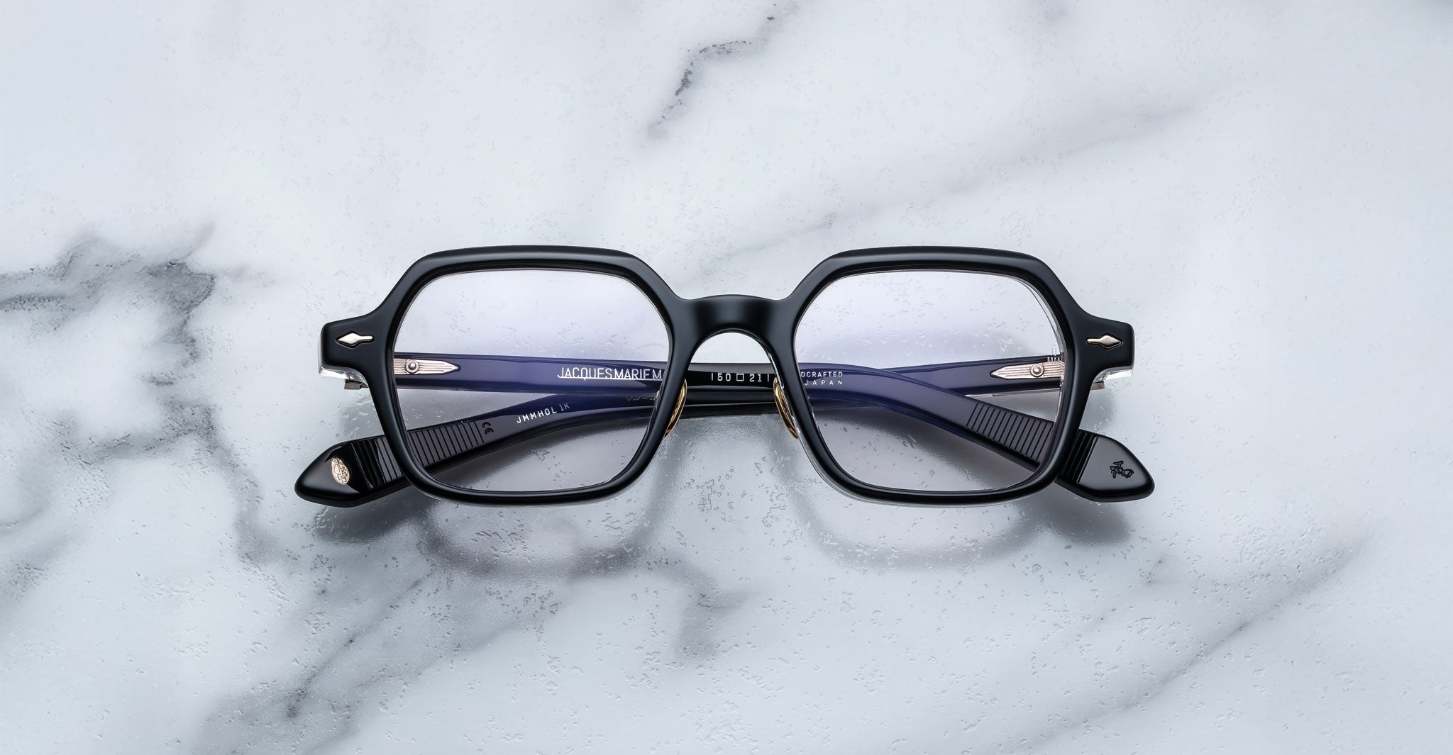 A pair of Jacques Marie Mage Holden black, square-framed eyeglasses with clear lenses rests on a white marble surface. The folded arms are visible through the lenses.