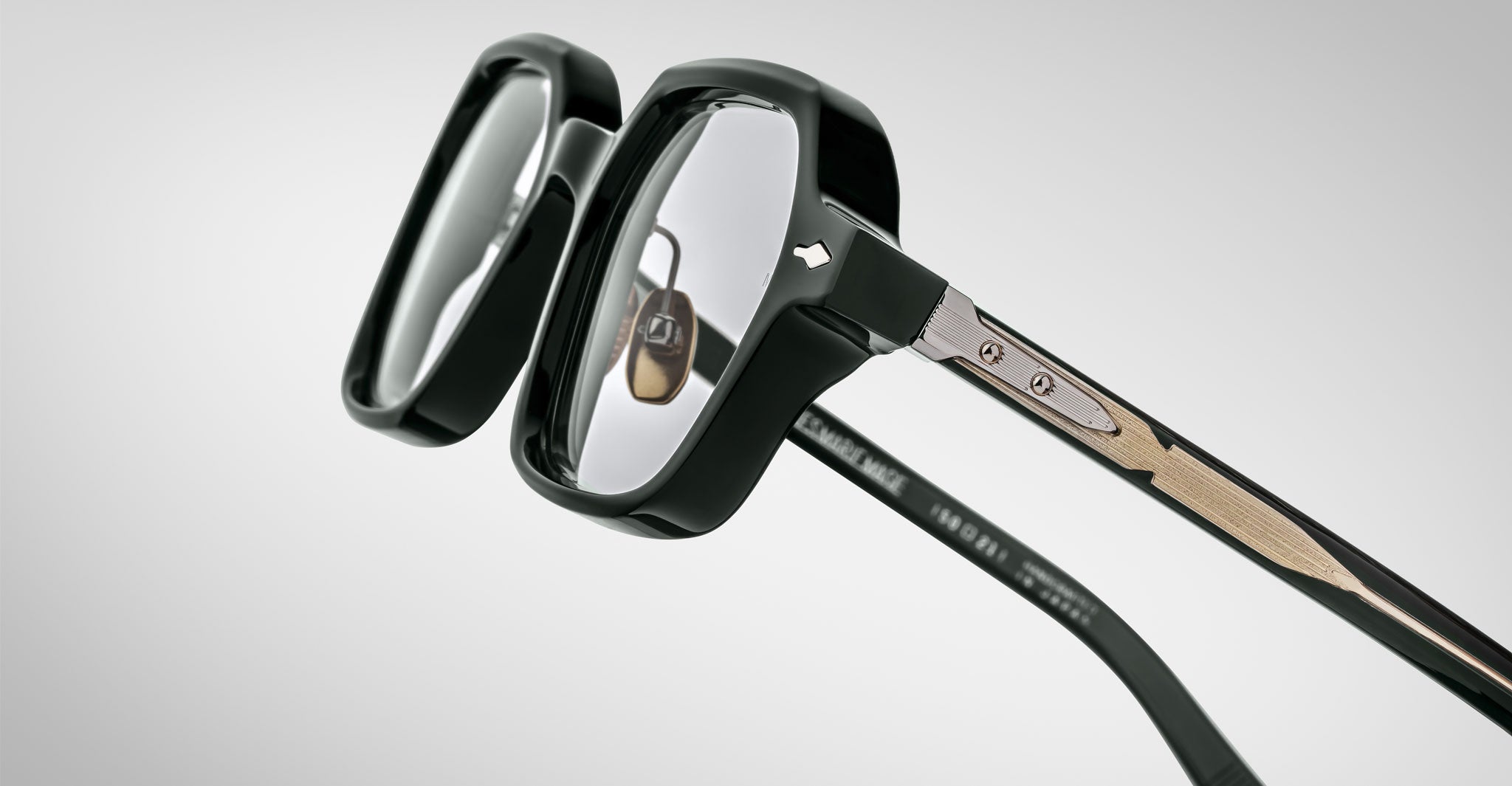 Close-up of Jacques Marie Mage Holden eyeglasses featuring thick black rims, clear lenses, and detailed metal and wood temple accents against a light gray background.