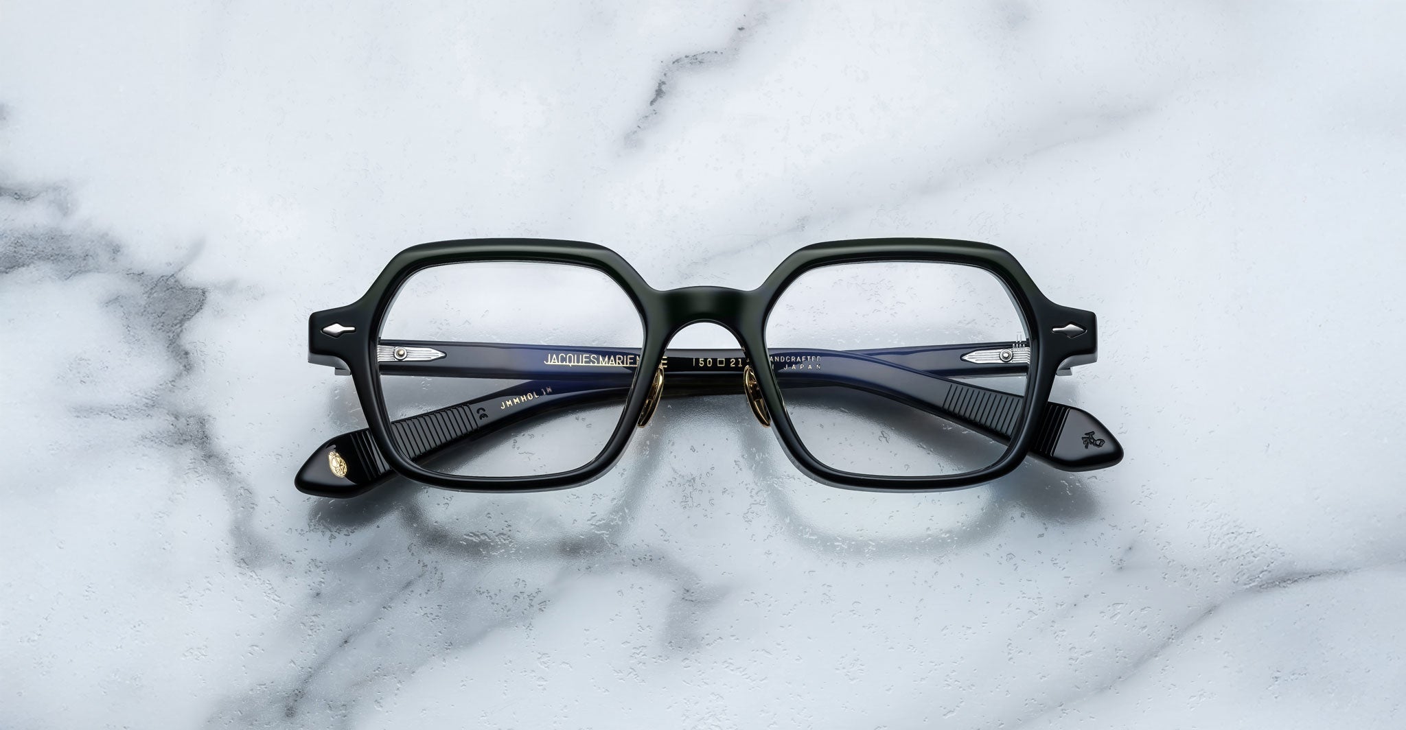 A pair of Jacques Marie Mage Holden black, square-rimmed eyeglasses with metal hinges rests on a white marble surface.