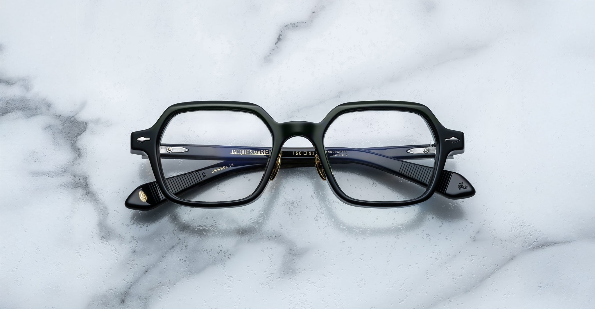 A pair of Jacques Marie Mage Holden black, square-rimmed eyeglasses with metal hinges rests on a white marble surface.