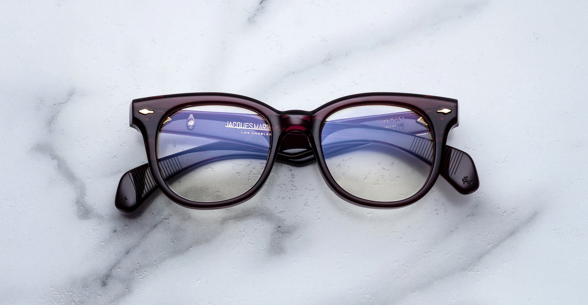 A pair of Jacques Marie Mage Hortense eyeglasses with dark rectangular frames rests on a white marble surface, their lenses reflecting light with a subtle blue tint.