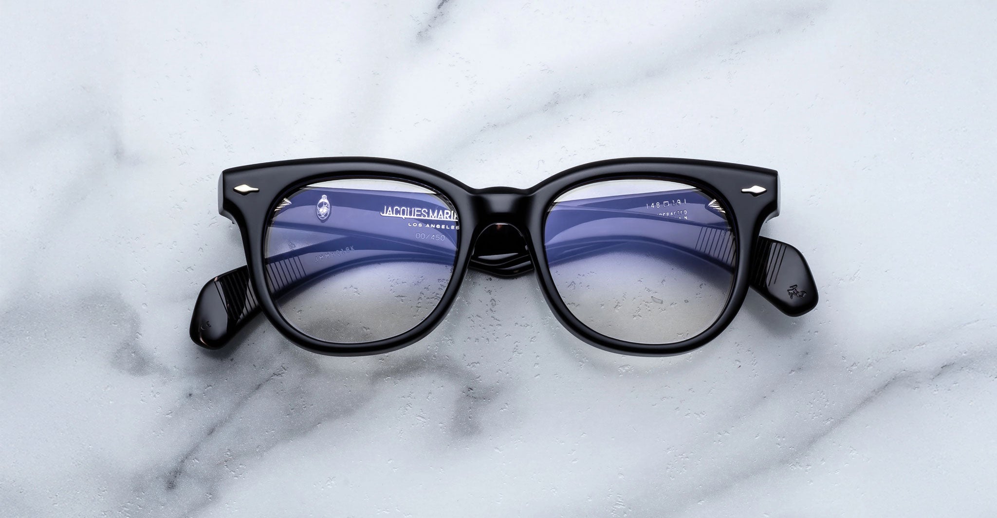 The Jacques Marie Mage Hortense eyeglasses, featuring black frames and slightly tinted lenses, are displayed on a white marble surface.