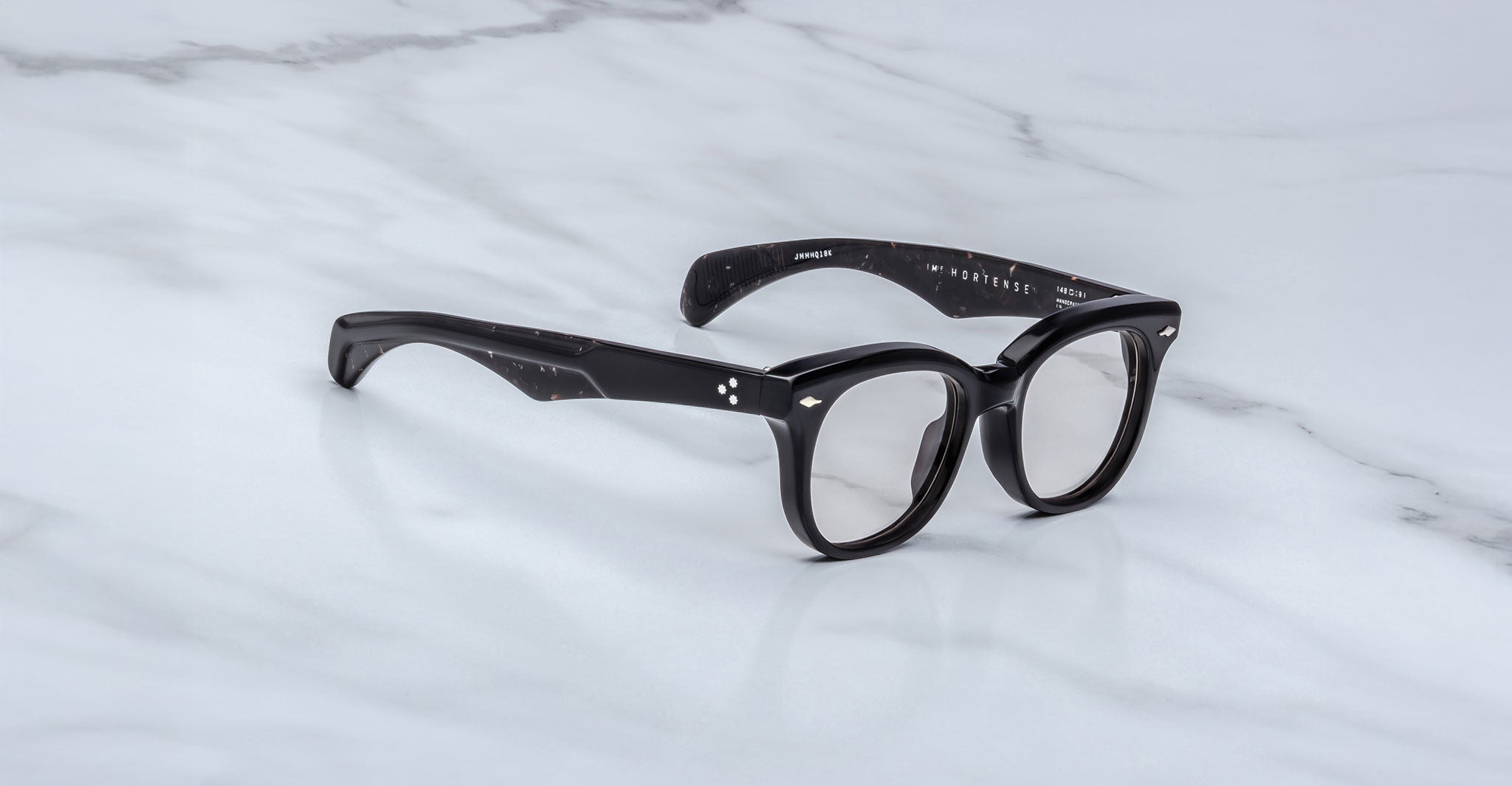 The Jacques Marie Mage Hortense eyeglasses feature black, thick round frames with subtle white speckles on the arms and silver accents by the hinges, displayed on a white marble surface.