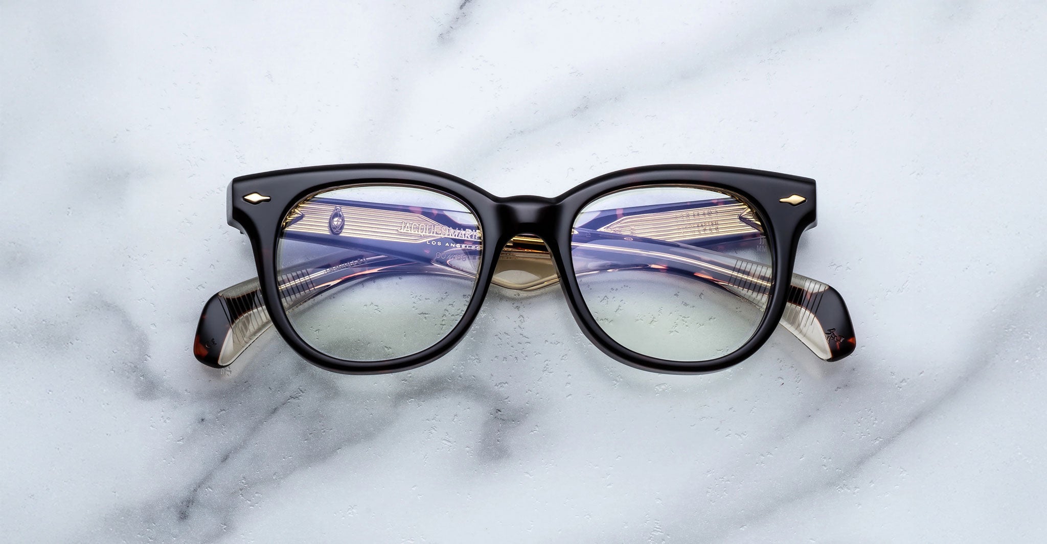 The Jacques Marie Mage Hortense eyeglasses, featuring black rims and round lenses with a subtle reflection, rest on a white marble surface; their temple tips display a striped pattern of dark and light hues.
