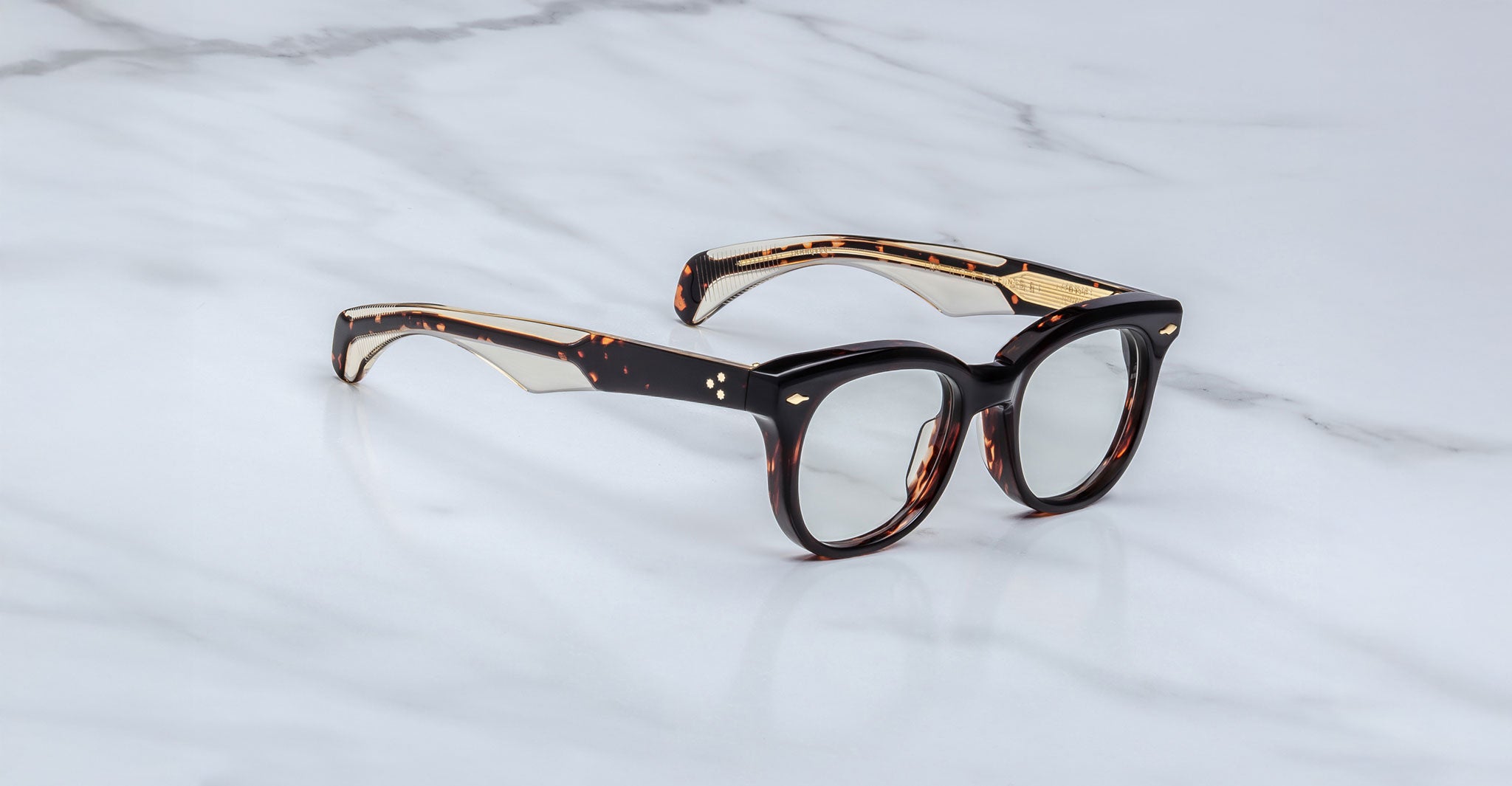 The Jacques Marie Mage Hortense features thick rims, a brown tortoiseshell pattern, and clear lenses, shown resting on a white marble surface.