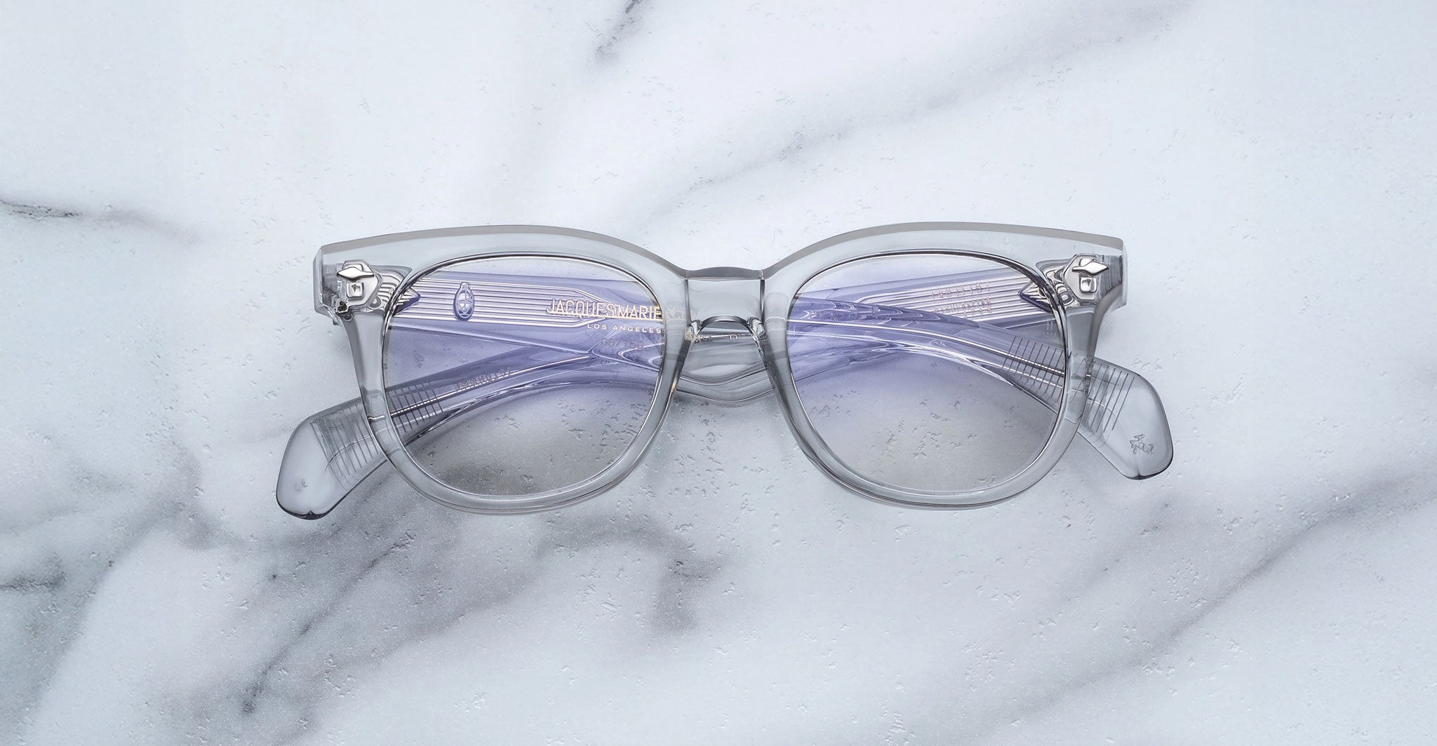 The Jacques Marie Mage Hortense glasses feature clear frames and lightly tinted purple lenses, set in a modern rectangular design with visible hinge details, displayed on a white marble surface.