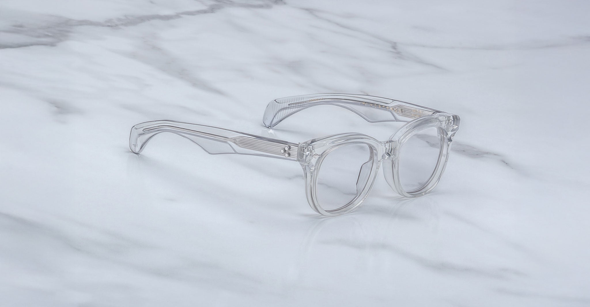 The Jacques Marie Mage Hortense features clear, thick-framed round lenses and is displayed on a white marble surface.