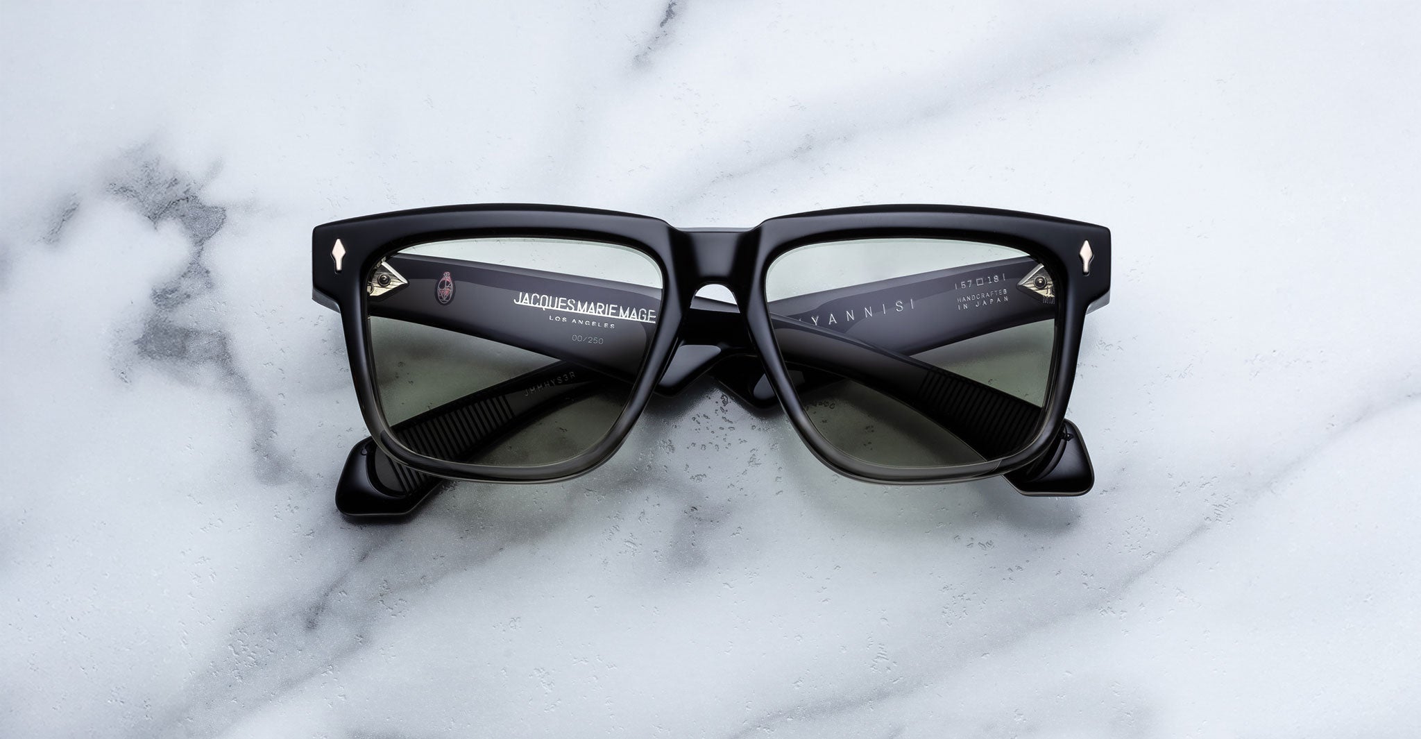 Jacques Marie Mage Hyannis black square-framed sunglasses with dark lenses rest on a white marble surface featuring subtle grey veining.