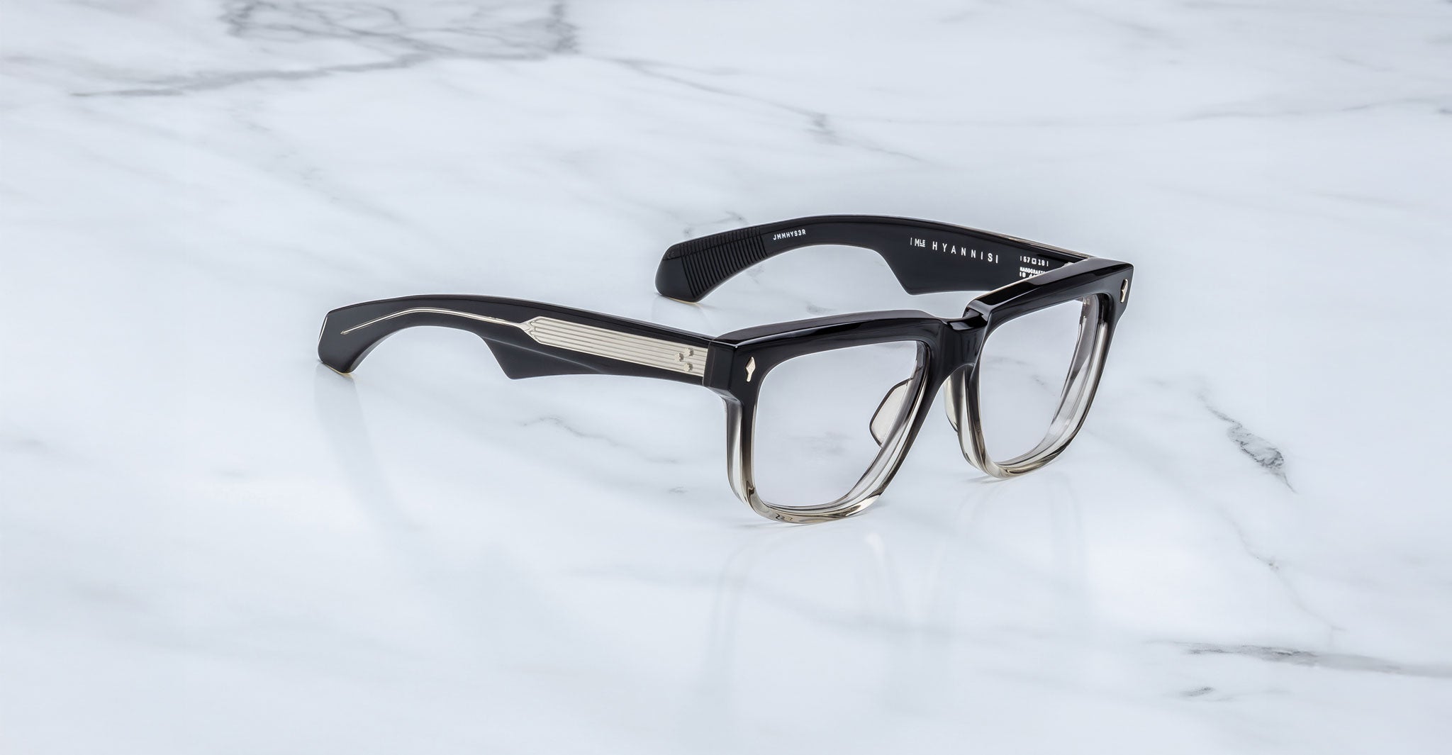 Jacques Marie Mage Hyannis rectangular eyeglass frames in a black-to-clear gradient with thick design and gold accents on the arms, shown on a white marble surface.