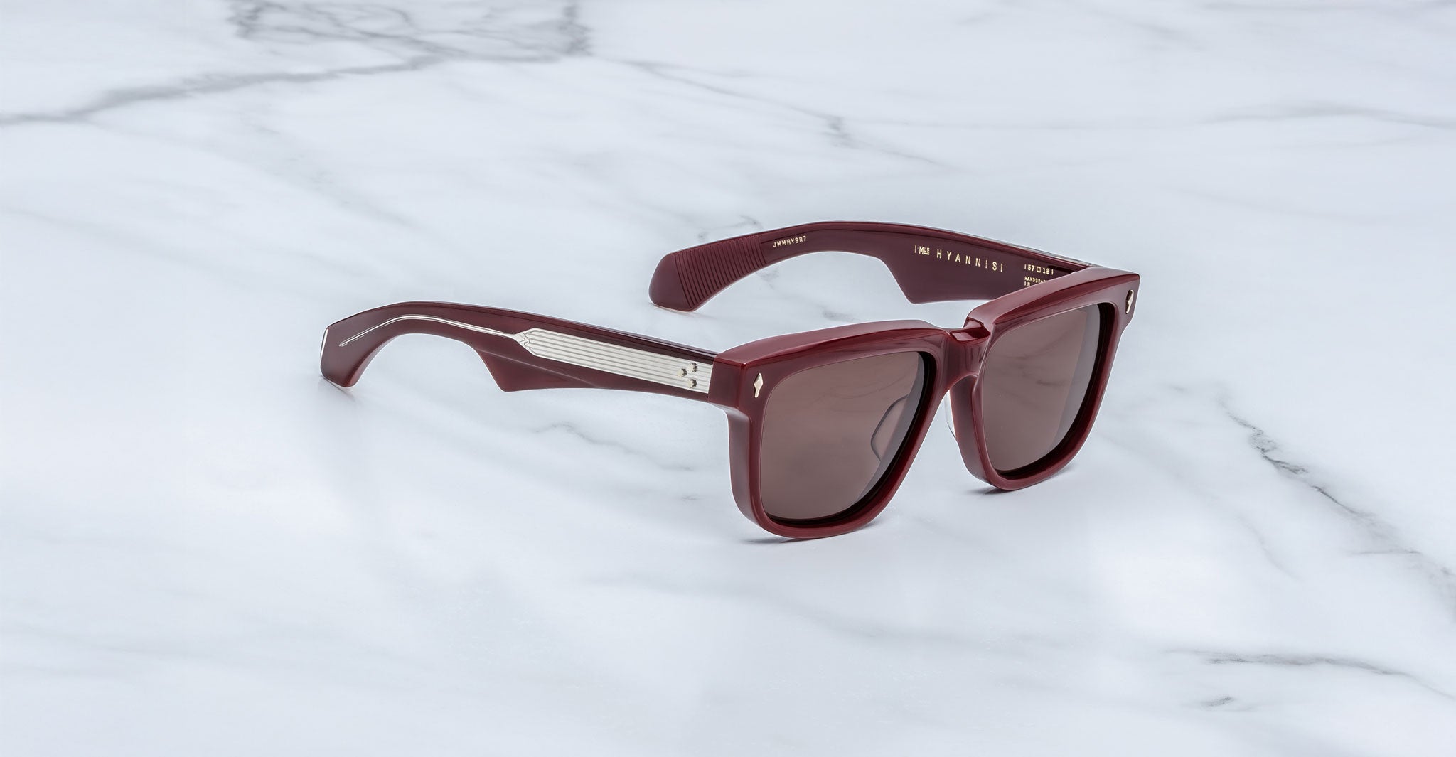 A pair of stylish Jacques Marie Mage Hyannis limited-edition maroon sunglasses with thick frames and dark lenses, displayed on white marble. The temples have metallic accents near the hinges.