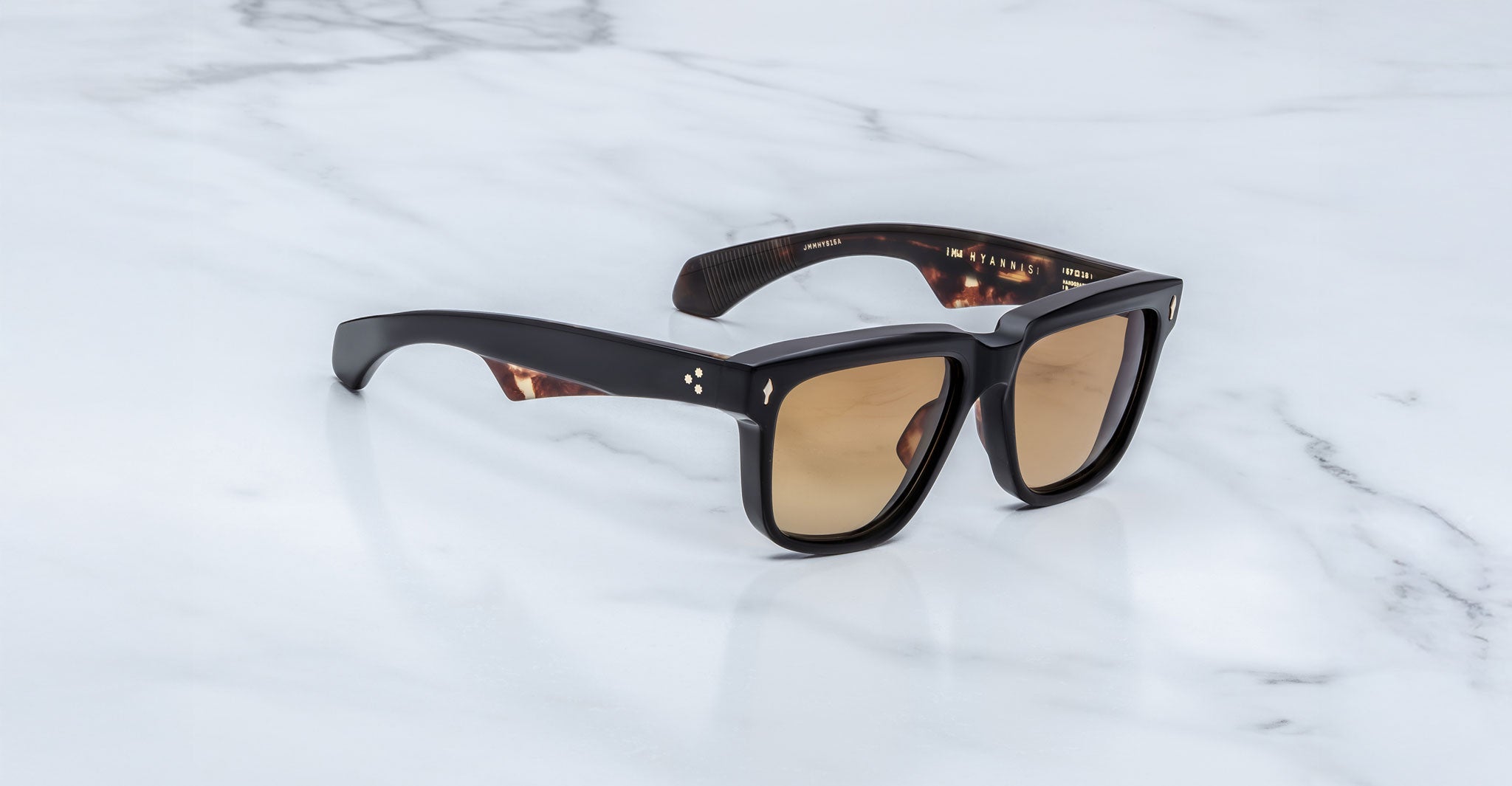A pair of Jacques Marie Mage Hyannis black, thick-framed sunglasses with brown-tinted lenses rests on a white marble surface, highlighting their exclusive limited-edition appeal.