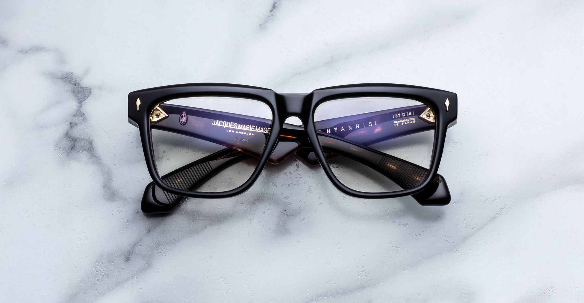 A pair of Jacques Marie Mage Hyannis black rectangular eyeglass frames with clear lenses rests on a white marble surface, showing text on the inside of the left arm.