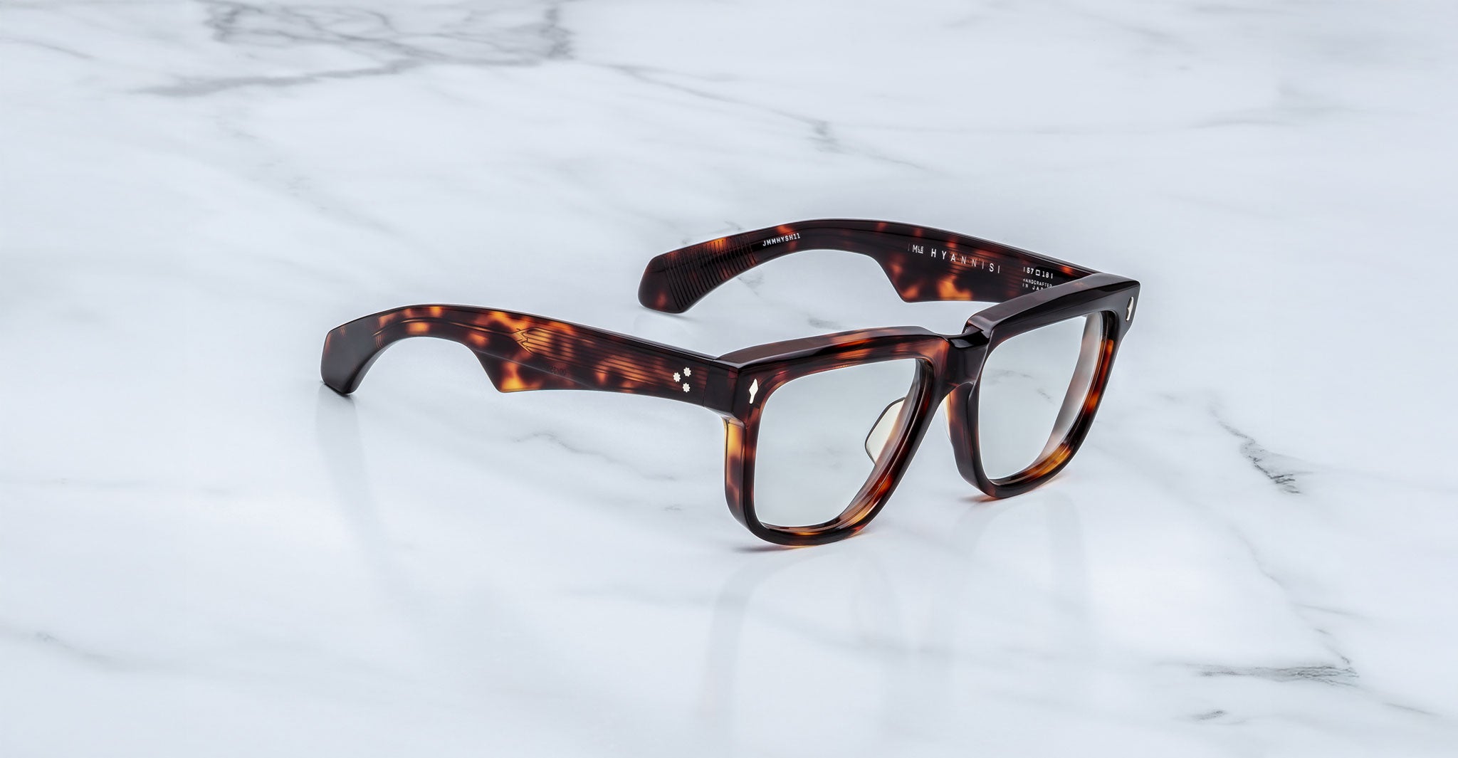 A pair of Jacques Marie Mage Hyannis eyeglasses with thick, rectangular tortoiseshell frames rests elegantly on a white marble surface.