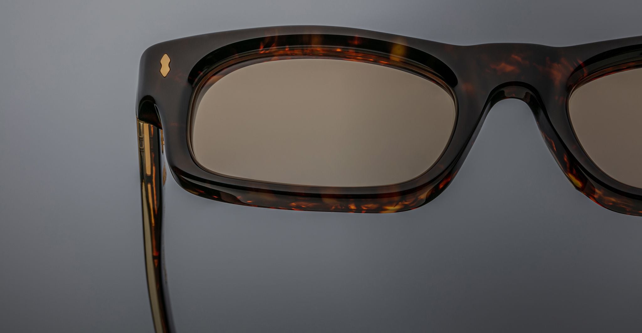Close-up of the right side of Jacques Marie Mage Initials rectangular tortoiseshell micro-frame sunglasses with brown-tinted lenses, set against a gray background.