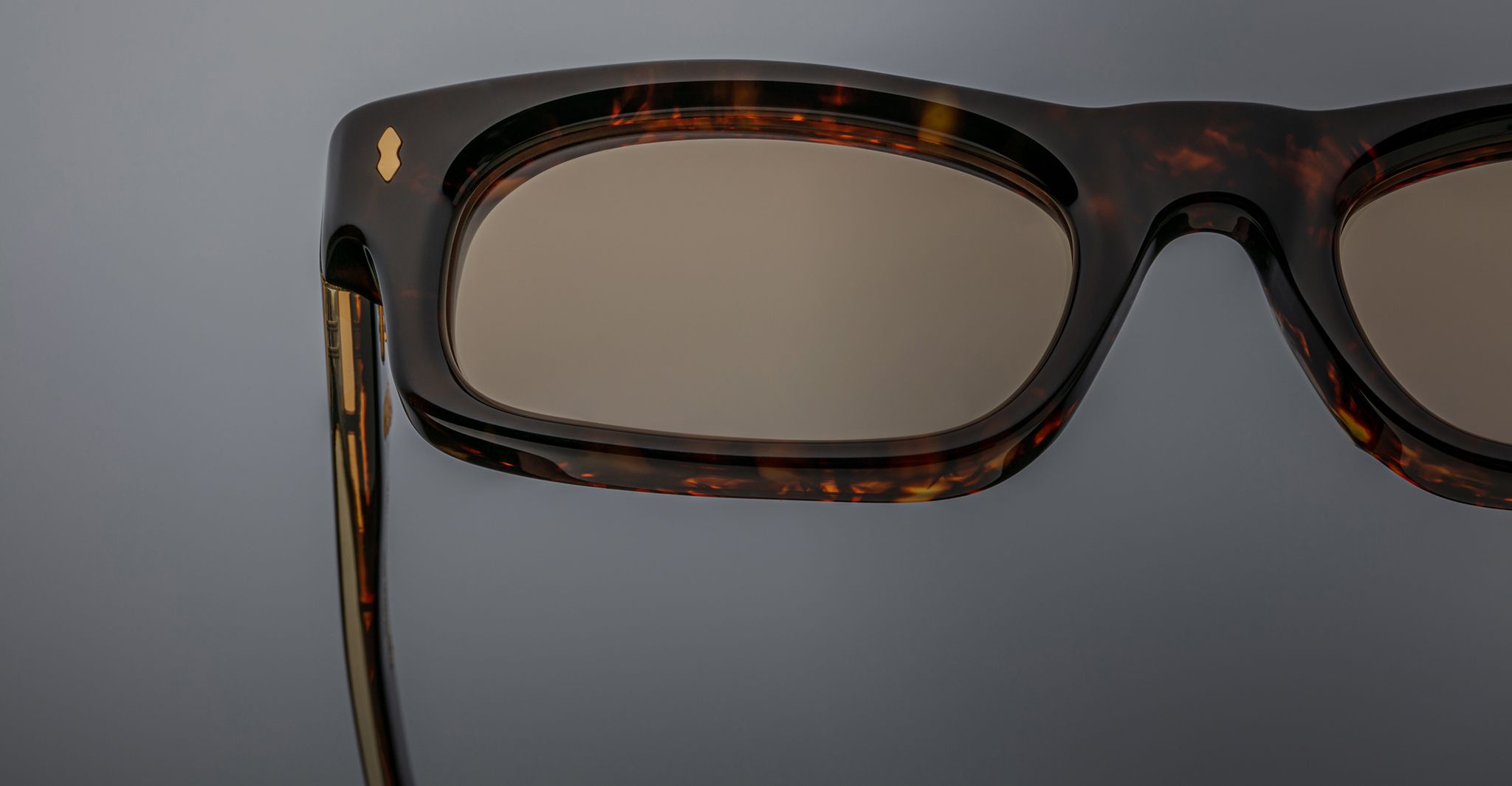 Close-up of the right side of Jacques Marie Mage Initials rectangular tortoiseshell micro-frame sunglasses with brown-tinted lenses, set against a gray background.