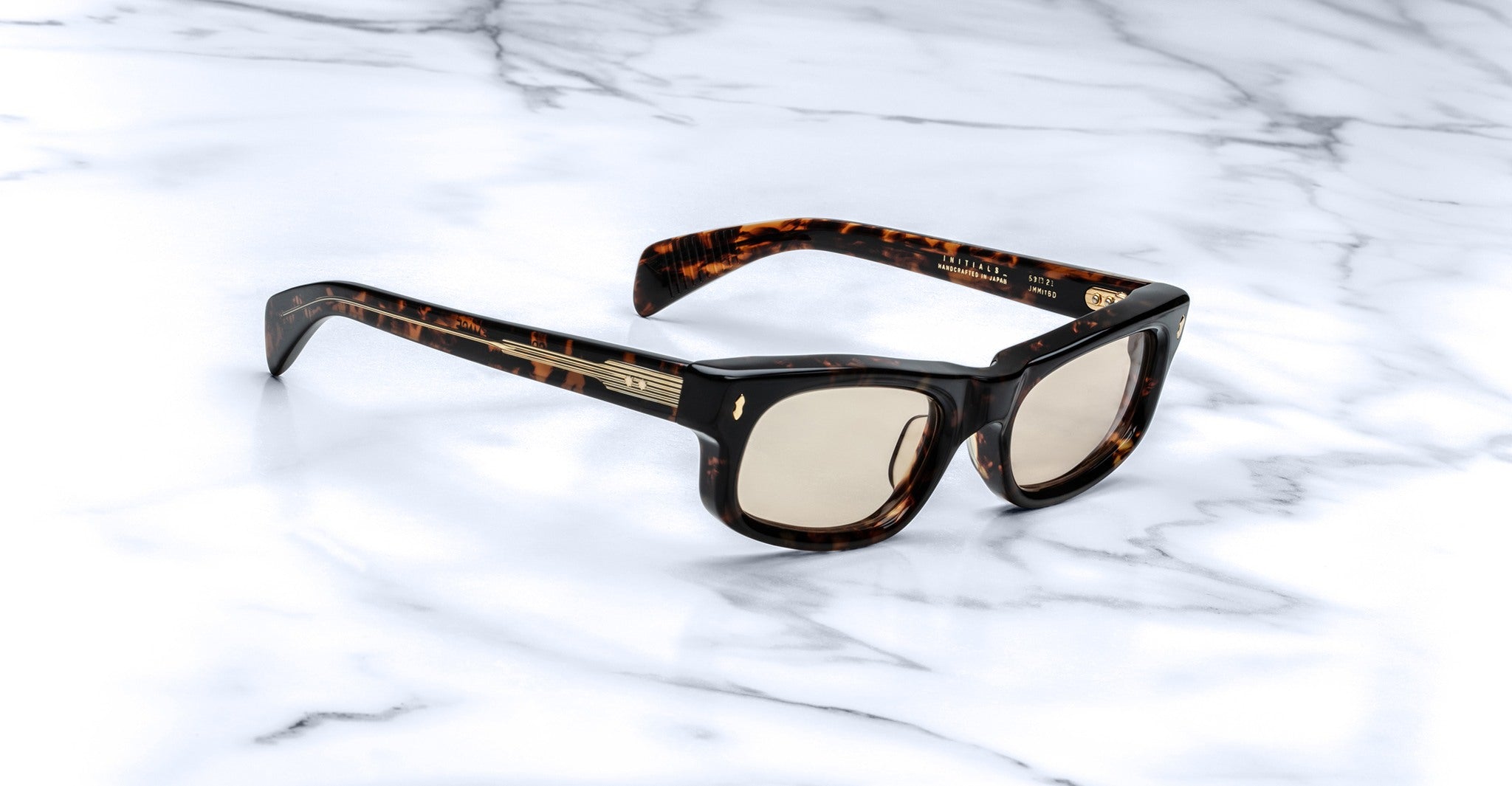 Jacques Marie Mage Initials tortoiseshell rectangular micro-frame sunglasses with light brown lenses rest on a white marble surface.