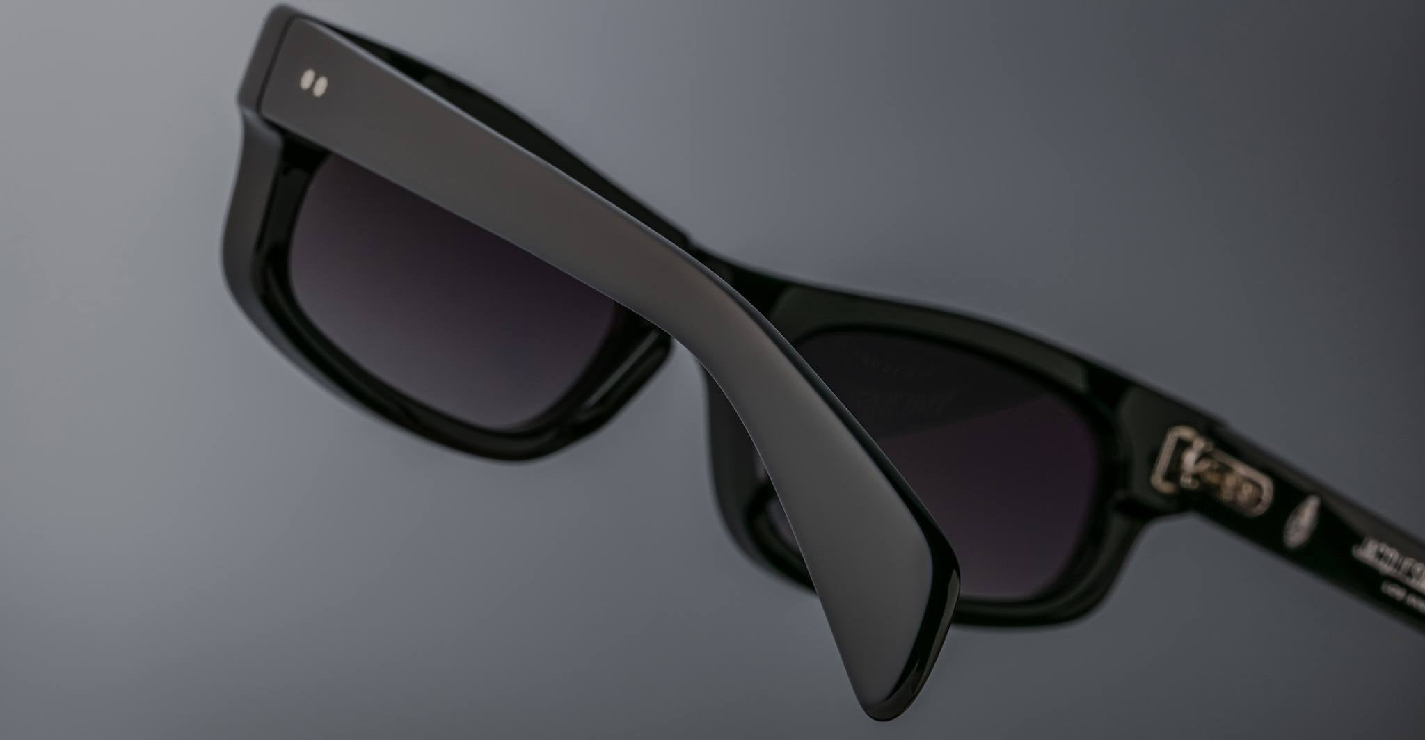 Close-up of Jacques Marie Mage Initials black rectangular sunglasses with dark lenses, set against a plain grey background. These limited-edition frames showcase the bold style and refined craftsmanship of Jacques Marie Mage.