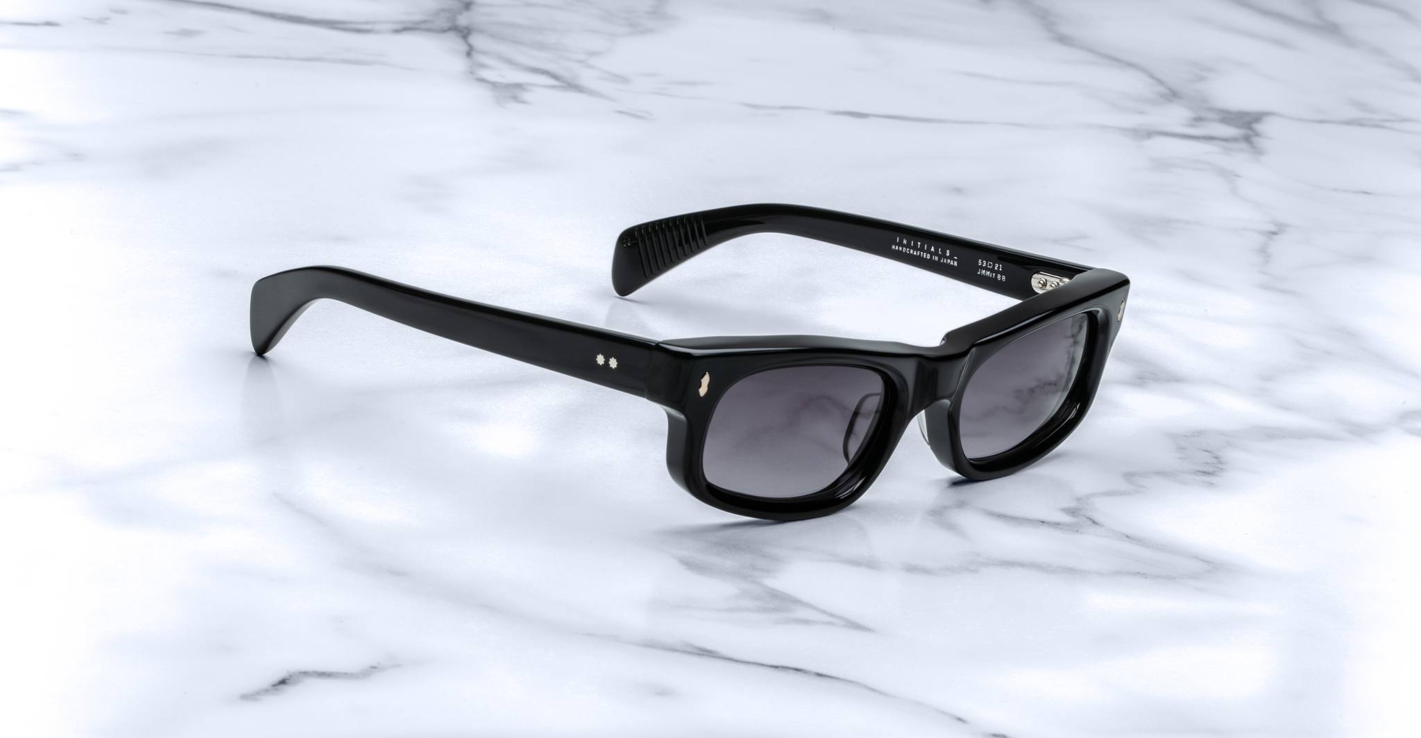 The Jacques Marie Mage Initials black rectangular micro-frame sunglasses with dark lenses rest on a white marble surface.