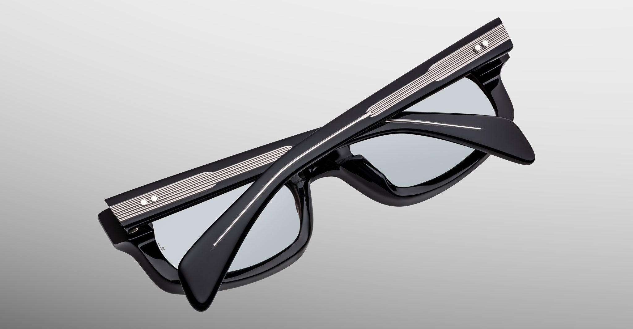 A pair of Jacques Marie Mage Initials sunglasses, black frames with metallic accents, is folded against a light gray background.
