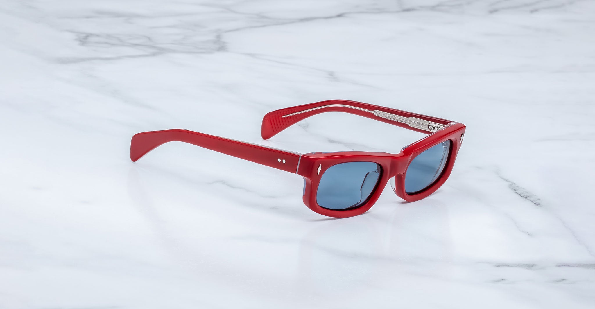 Jacques Marie Mage Initials sunglasses with red frames and dark lenses rest on a white marble surface, showcasing the brand's exclusive limited-edition style.