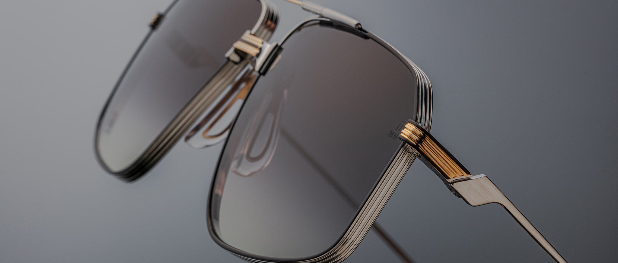 Close-up of Jacques Marie Mage Jagger sunglasses by Jacques Marie Mage, showcasing oversized aviator style with gold and silver beta titanium frames and rectangular gradient tinted lenses against a gray background.