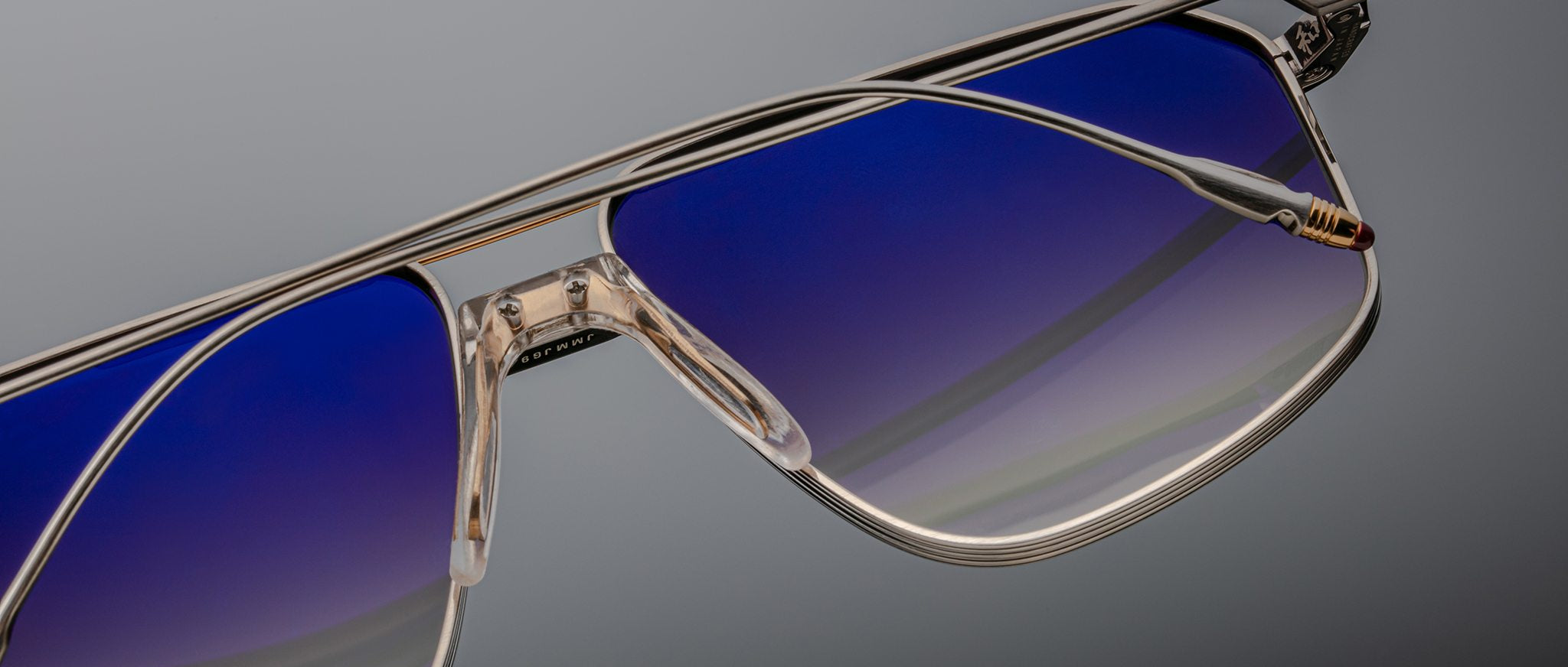 Close-up of Jacques Marie Mage Jagger oversized aviator sunglasses by Jacques Marie Mage, featuring beta titanium metallic frames and blue gradient lenses against a plain gray background.