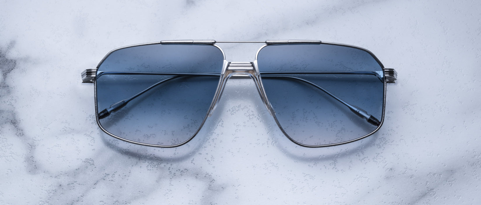 A pair of Jacques Marie Mage Jagger oversized aviator sunglasses with blue lenses and a sleek silver beta titanium frame by Jacques Marie Mage, displayed on a white marble surface.