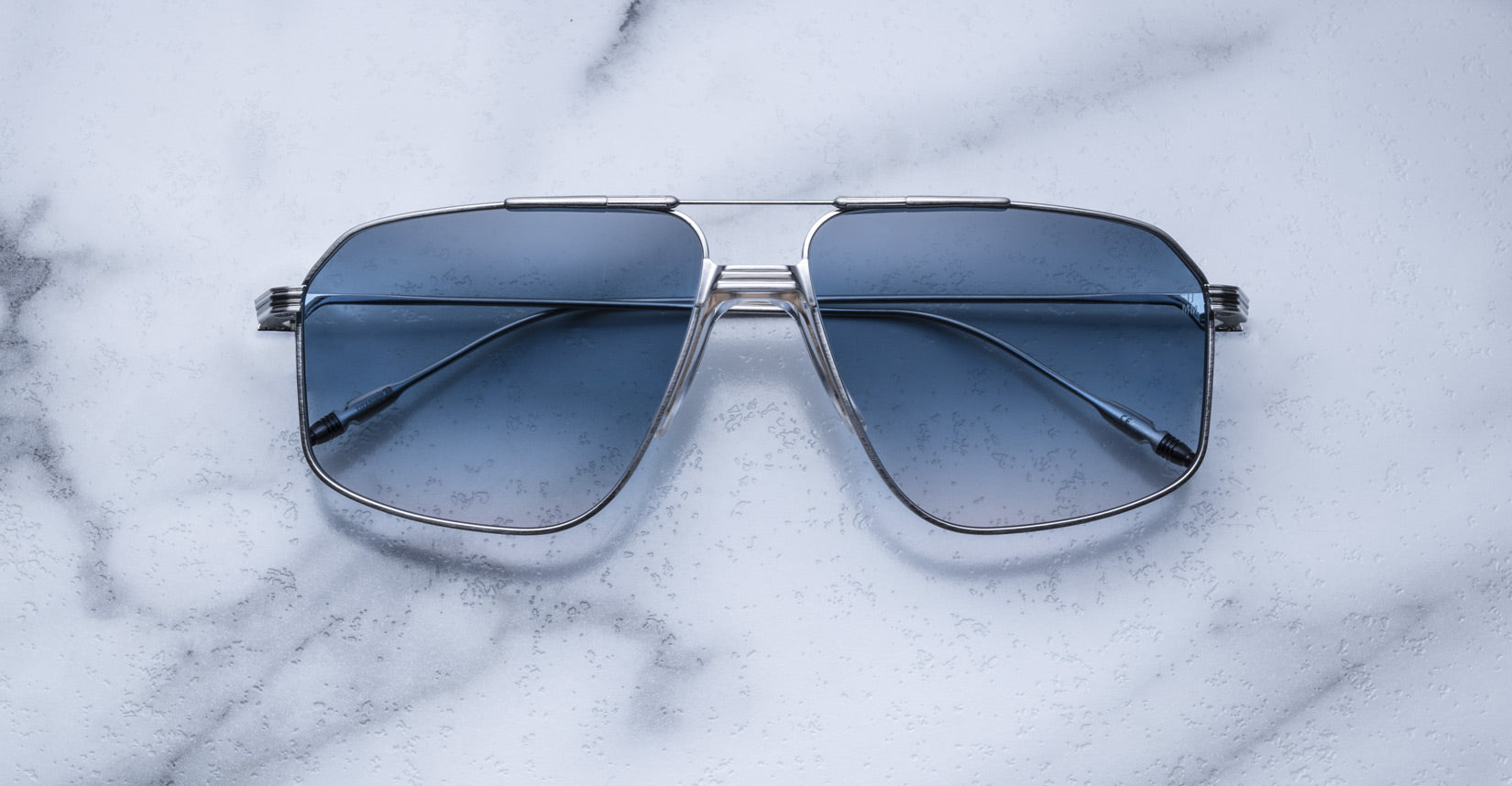 A pair of Jacques Marie Mage Jagger oversized aviator sunglasses with blue lenses and a sleek silver beta titanium frame by Jacques Marie Mage, displayed on a white marble surface.