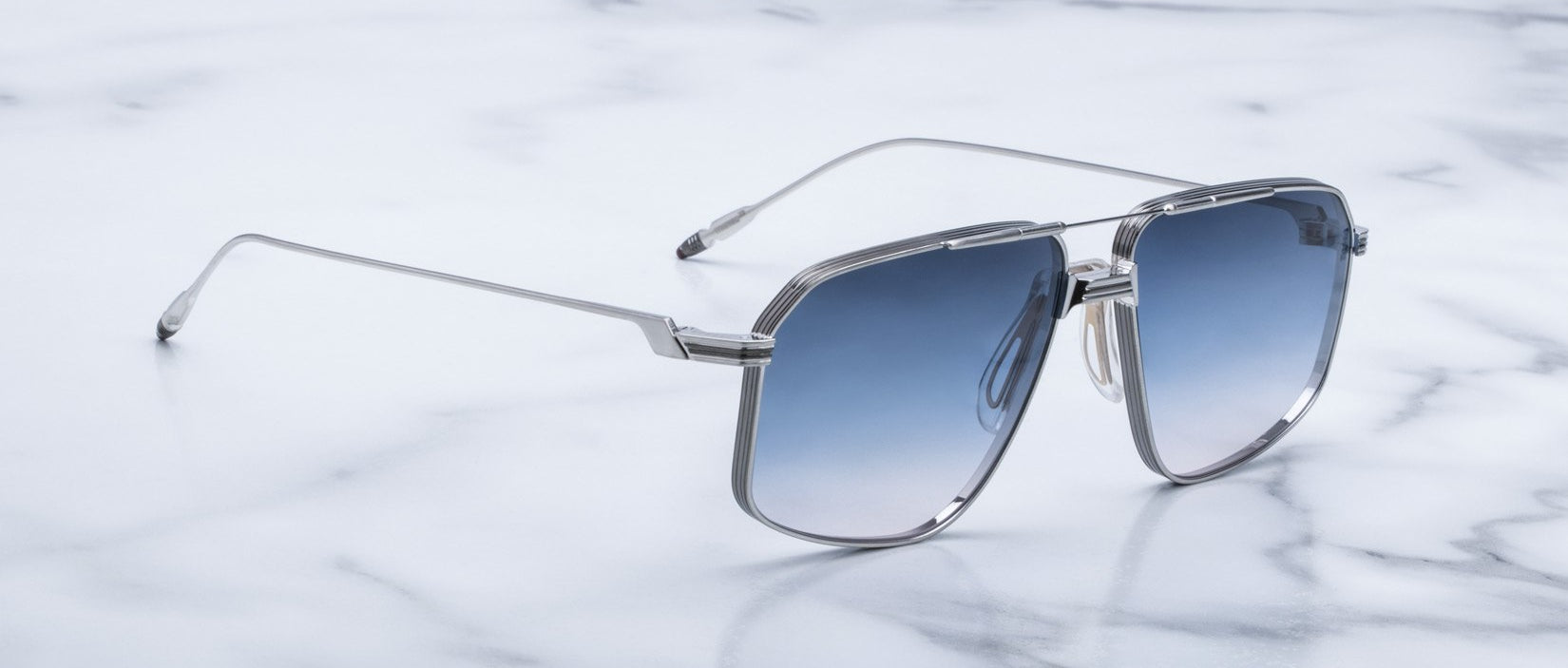 Jacques Marie Mage Jagger oversized aviator sunglasses with a silver frame and blue gradient lenses rest on white marble, channeling the iconic style of Jacques Marie Mage.