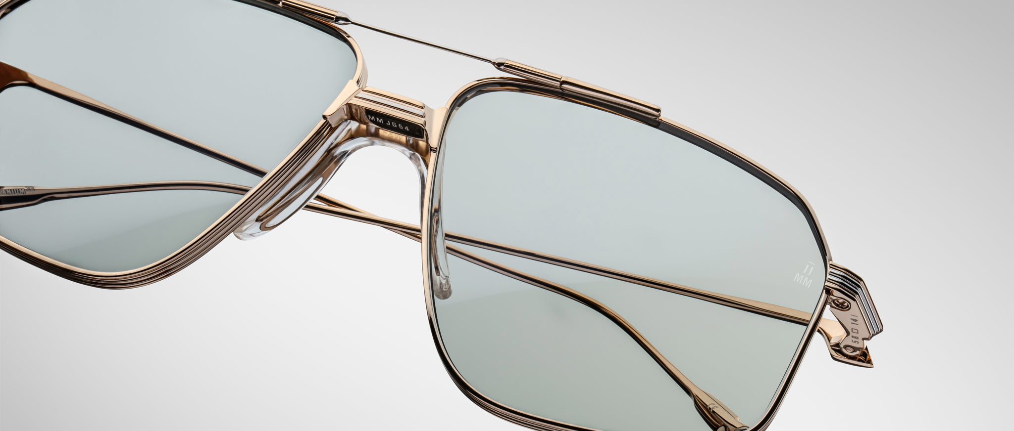 Close-up of the Jacques Marie Mage Jagger oversized aviator sunglasses by Jacques Marie Mage, with gold frames and light blue-tinted lenses set against a plain, gradient background.
