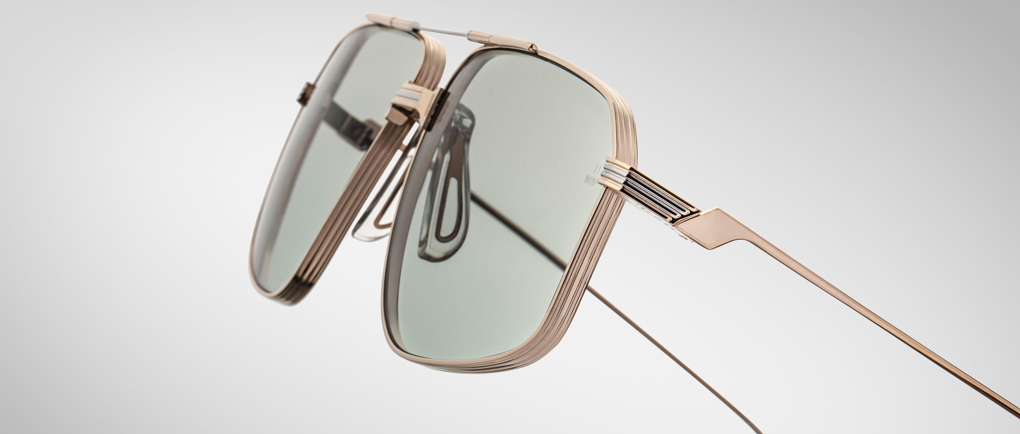 Close-up of Jacques Marie Mage Jagger sunglasses featuring gold beta titanium frames and rectangular green-tinted lenses by Jacques Marie Mage, displayed against a plain light gray background.