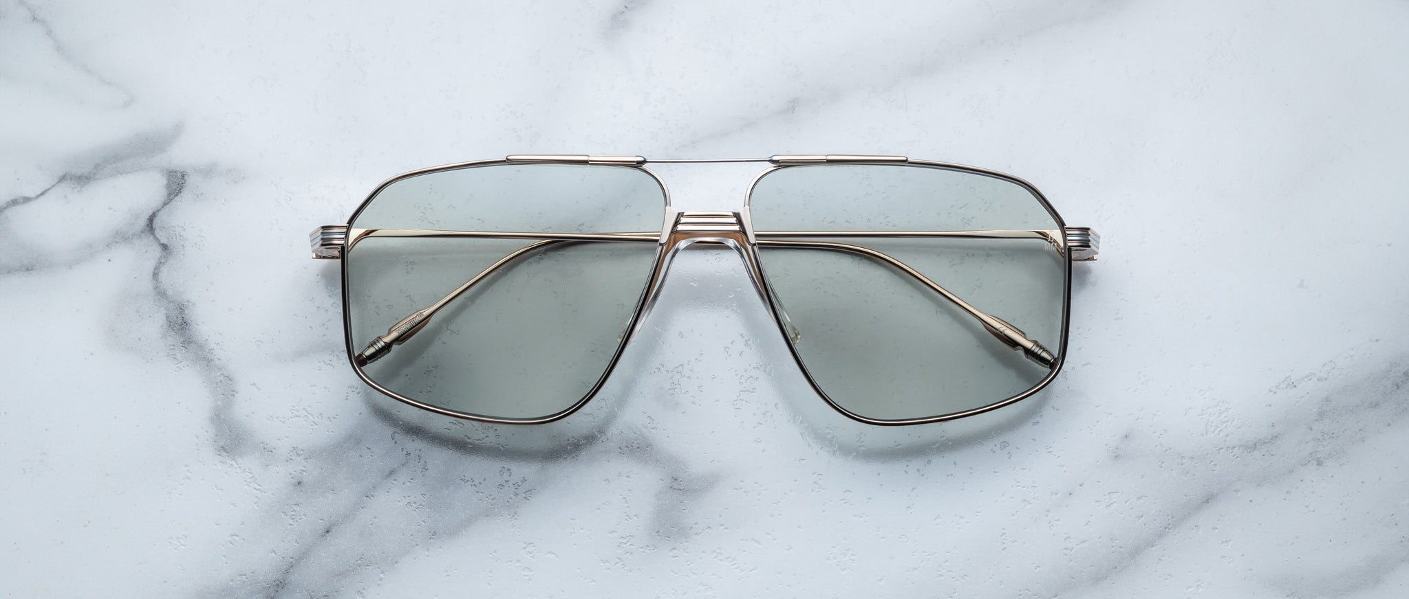 Jacques Marie Mage Jagger oversized aviator sunglasses by Jacques Marie Mage, featuring gold beta titanium frames and tinted lenses, are shown on a white marble surface.