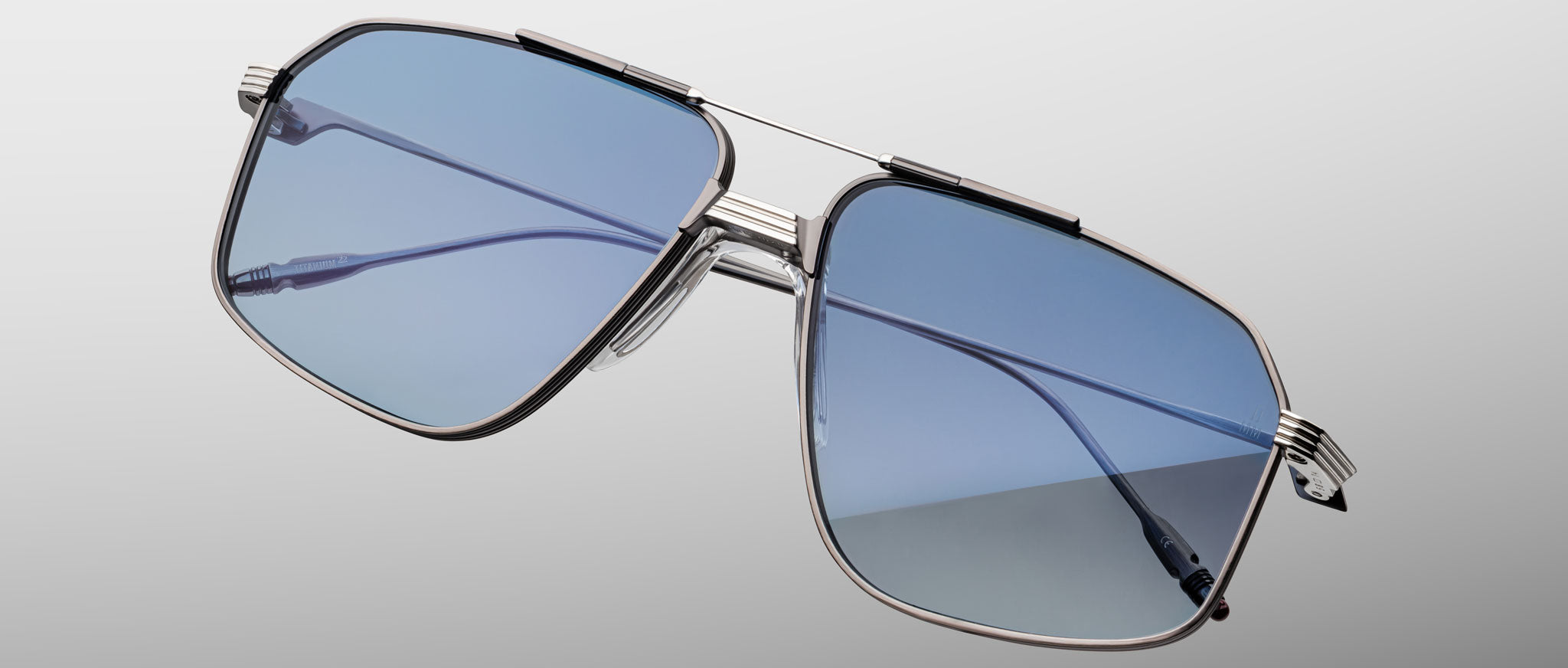 Close-up of Jacques Marie Mage Jagger oversized aviator sunglasses with blue-tinted lenses and metal frames, set against a plain gradient grey background.