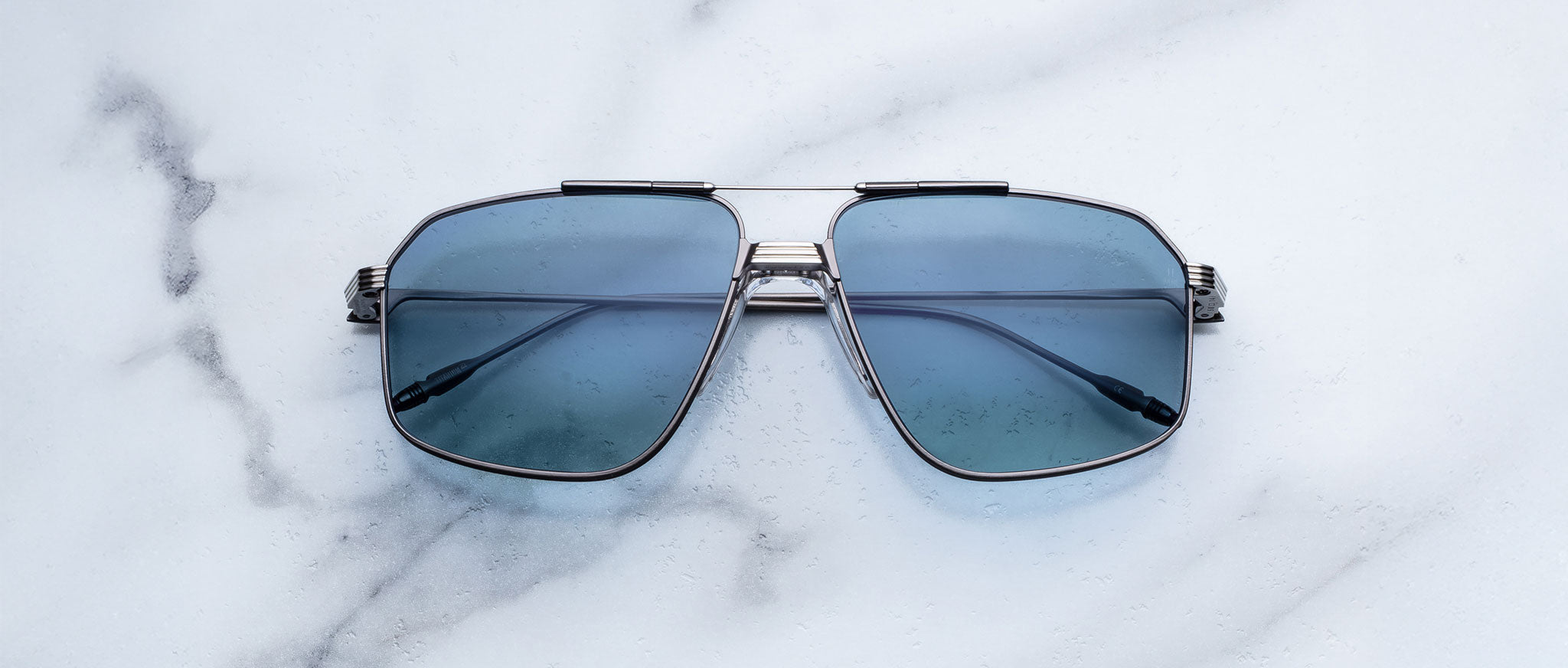 A pair of Jacques Marie Mage Jagger oversized aviator sunglasses with blue-tinted lenses and beta titanium frames by Jacques Marie Mage is displayed on a white marble surface.