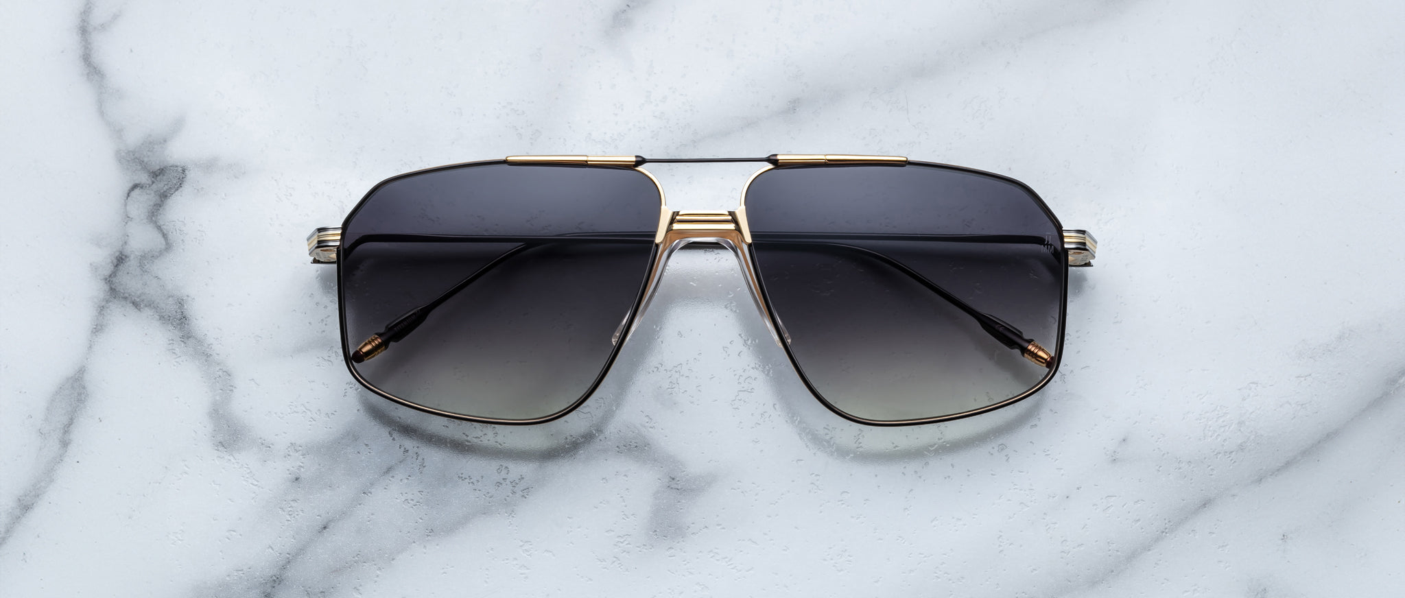 The Jacques Marie Mage Jagger sunglasses by Jacques Marie Mage, black oversized aviators with gold and beta titanium accents, are displayed on a white marble surface.