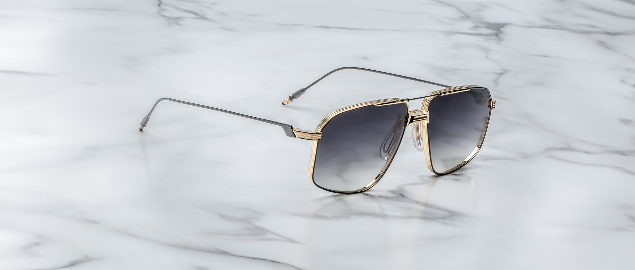 The Jacques Marie Mage Jagger sunglasses by Jacques Marie Mage feature oversized aviator frames with gold rims and dark lenses, exuding sophistication as they are elegantly displayed on a white marble surface.